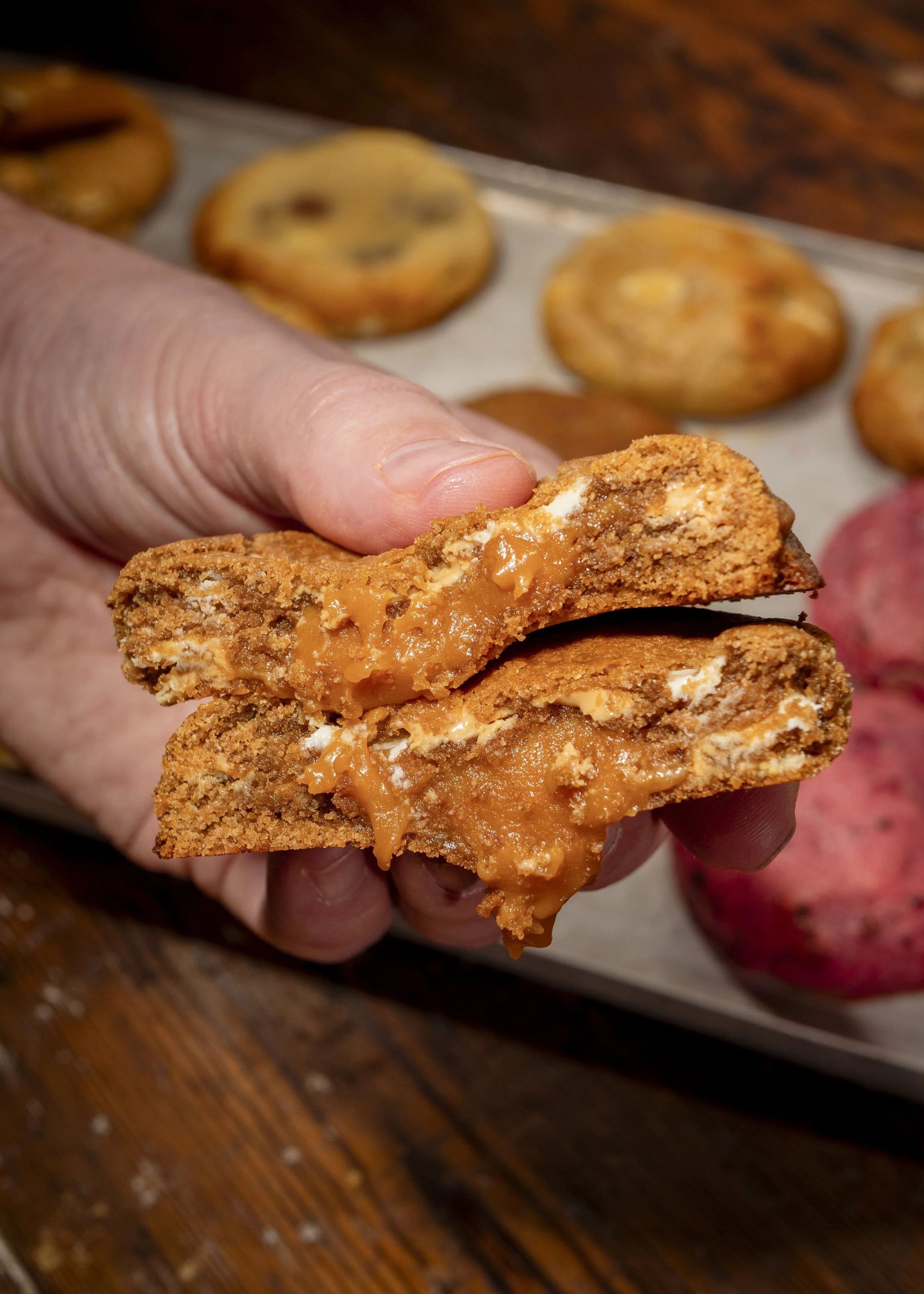 Hand holding a peanut butter cookie broken in half with melted peanut butter inside, with cookies and sweet potatoes in the background.