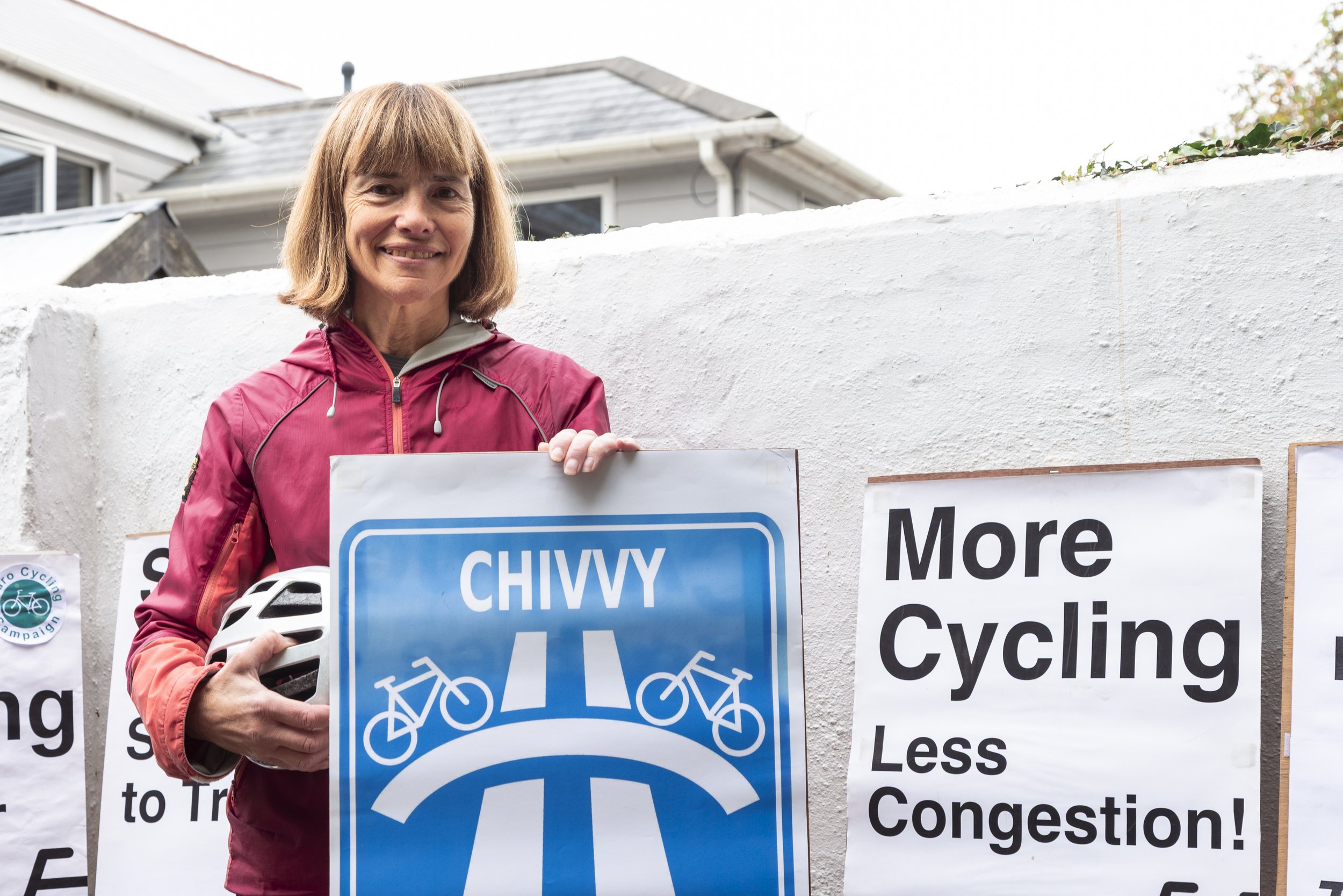 A woman standing outdoors in front of a white wall, holding a bike helmet and a sign that says 'Chivvy' with a bicycle lane graphic, with other signs promoting cycling and less congestion.