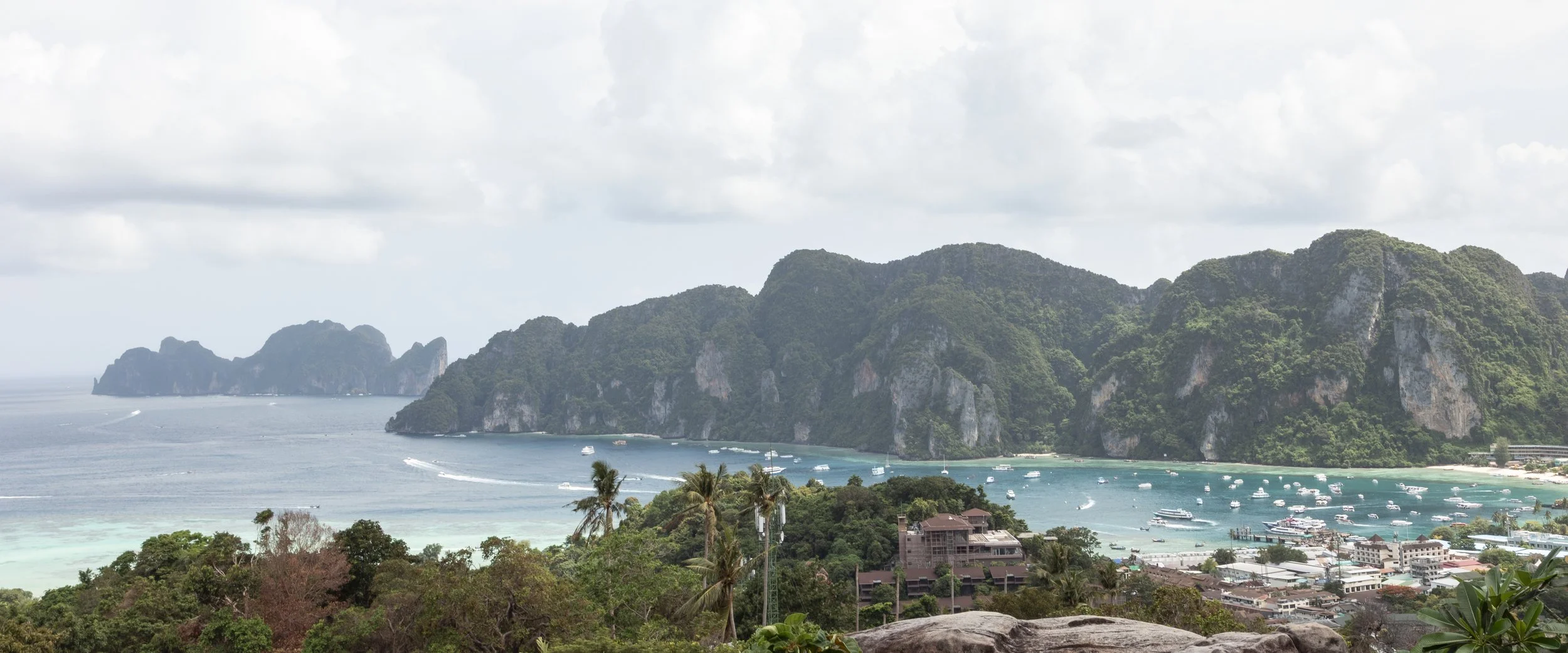 A coastal landscape with turquoise waters, lush green hills, and a small town with boats anchored near the shore, under a partly cloudy sky.