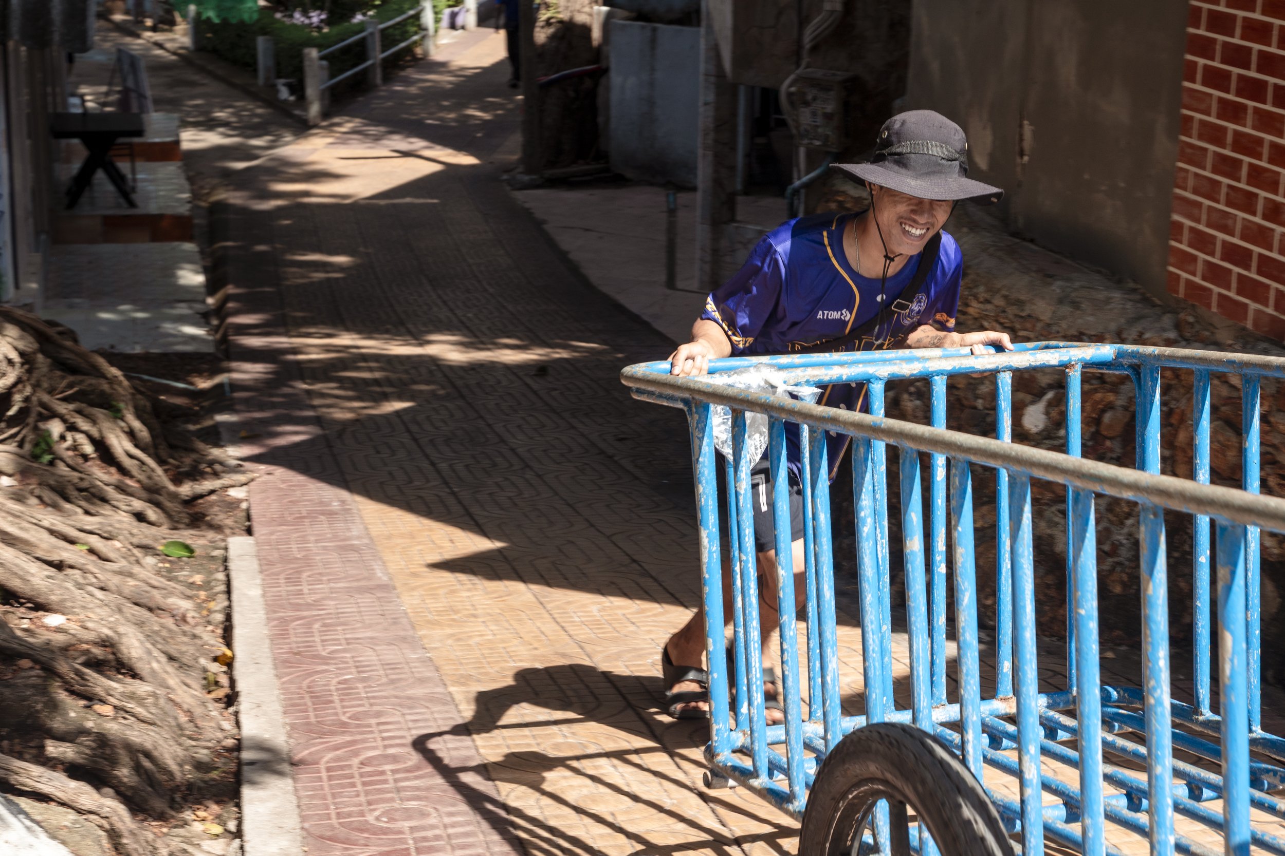 A man wearing a wide-brimmed hat and a blue sports jersey, pushing a blue cart on a sidewalk and smiling.