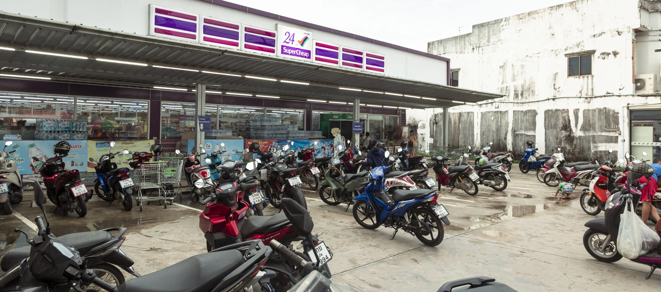 Parking lot outside a store with numerous motorcycles parked and some people around, with a store entrance and signage in the background.