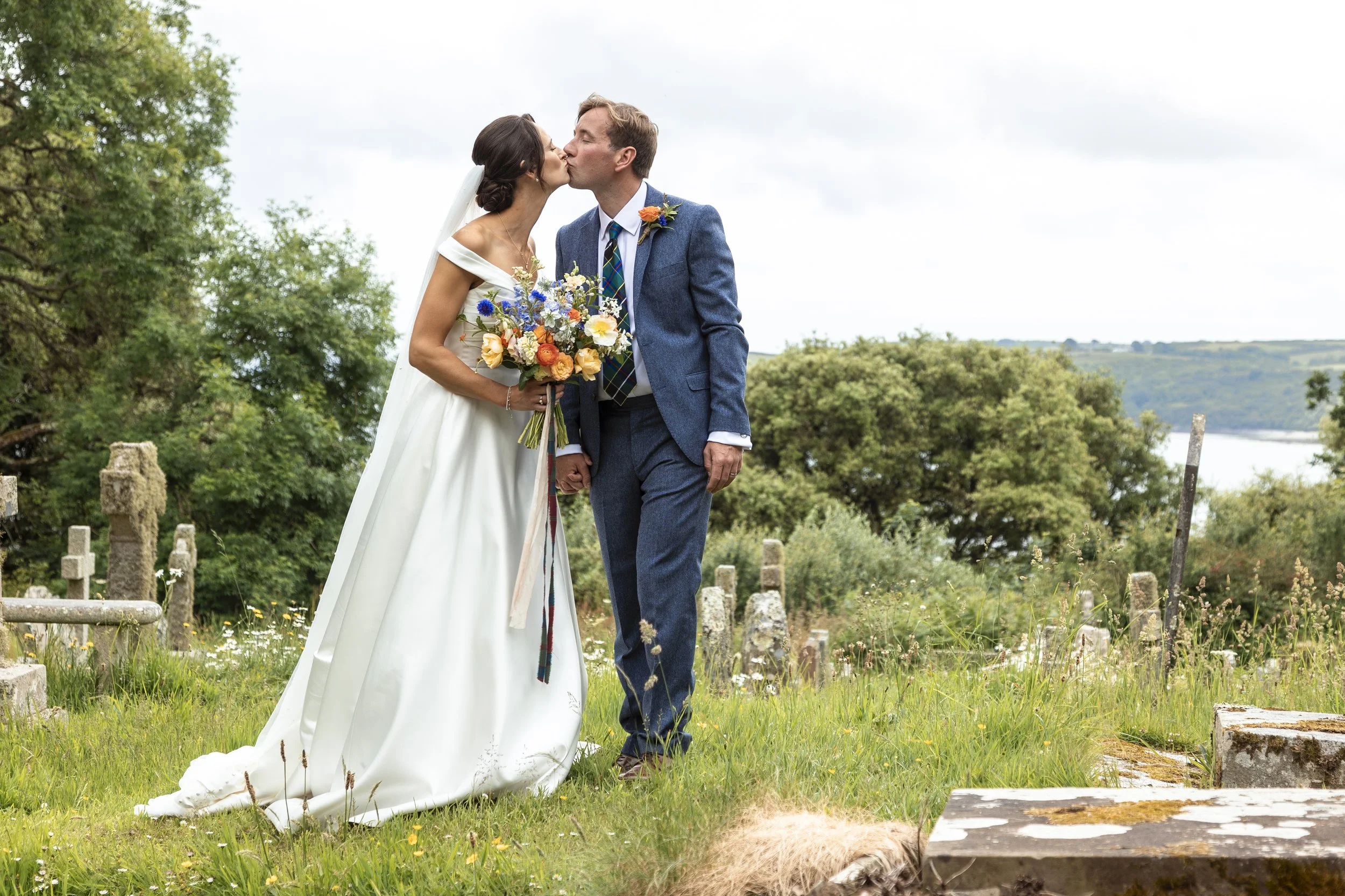 Bride and groom kissing outdoors, bride holding a colorful bouquet, grassy field with stone ruins, trees, and a lake in the background.