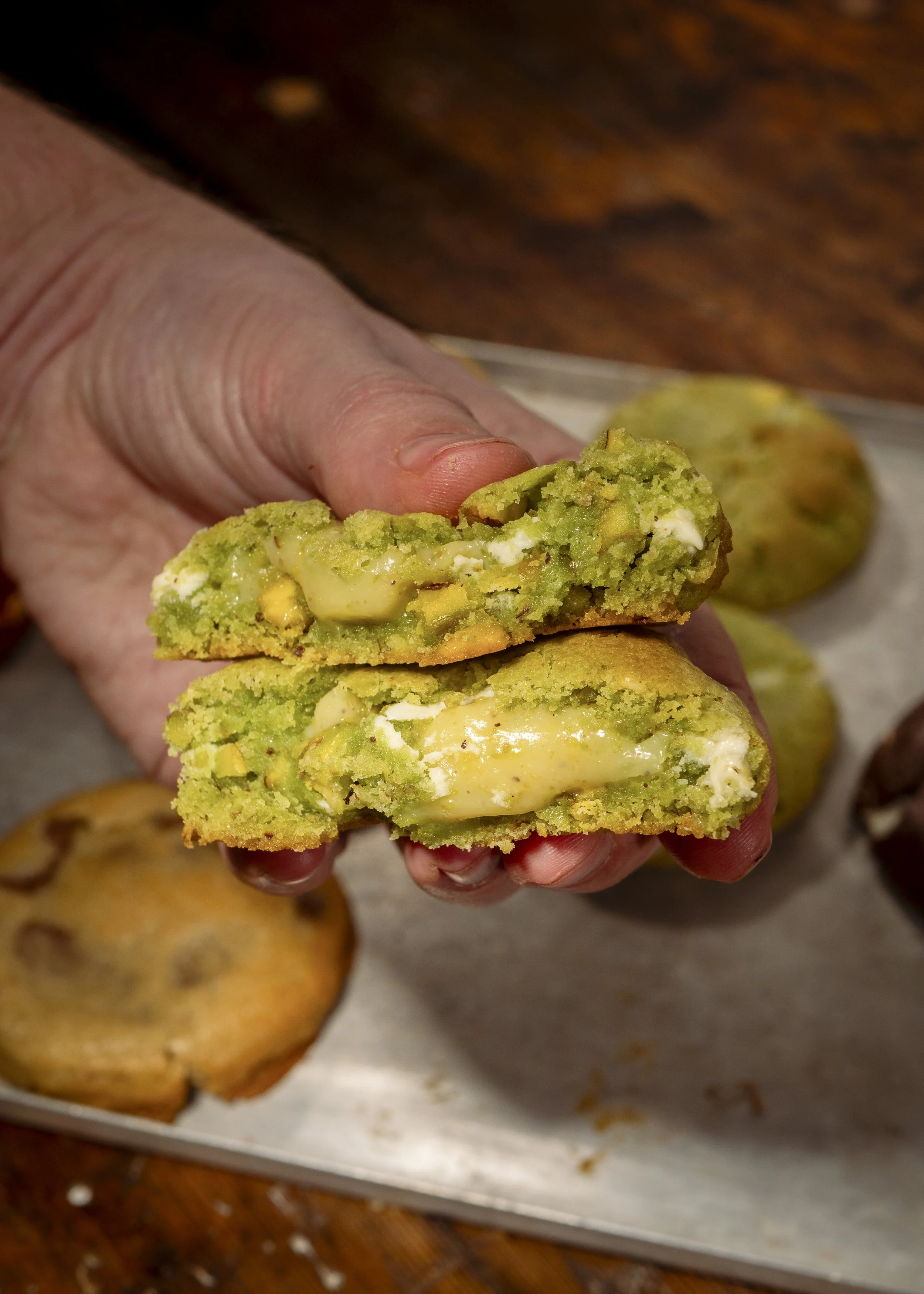 A hand holding a green cookie with white chocolate chips, showing a bite to reveal a creamy filling inside, with more cookies on a tray in the background.