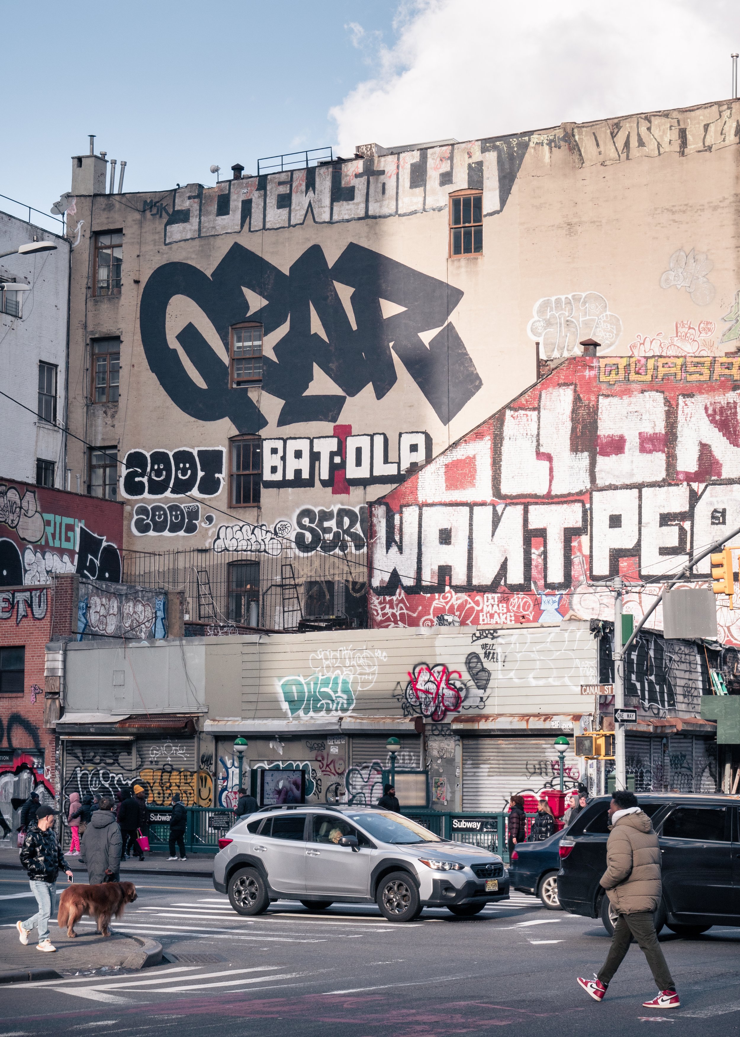 Street scene with pedestrians crossing and cars driving past a building covered in graffiti murals and tags.