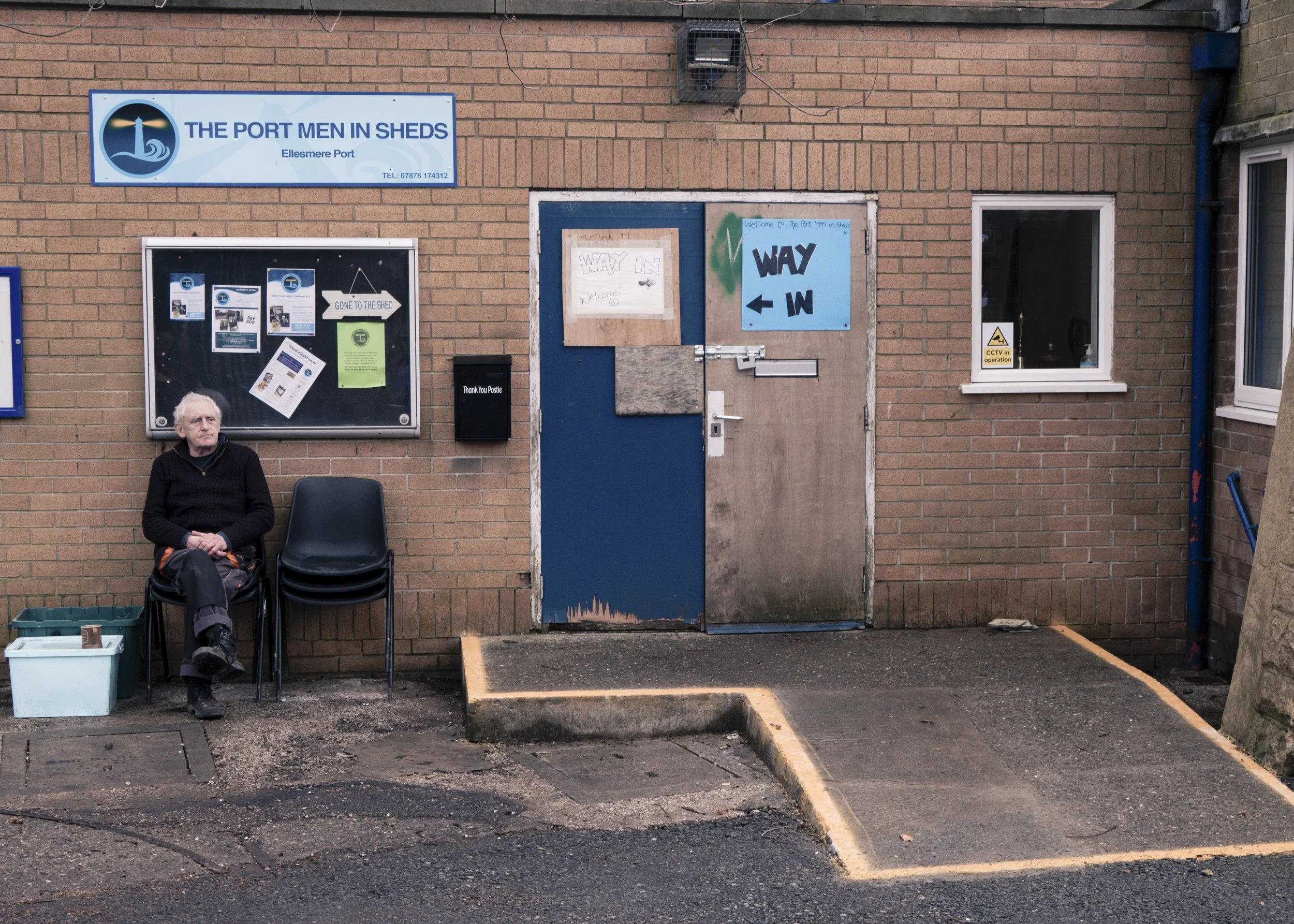A man with white hair sitting on a black plastic chair outside a brick building, next to a stack of black chairs. There is a blue and beige double door with signs and boards, one sign says 'Way In' with arrows. A window with a yellow 'CCTV in operati