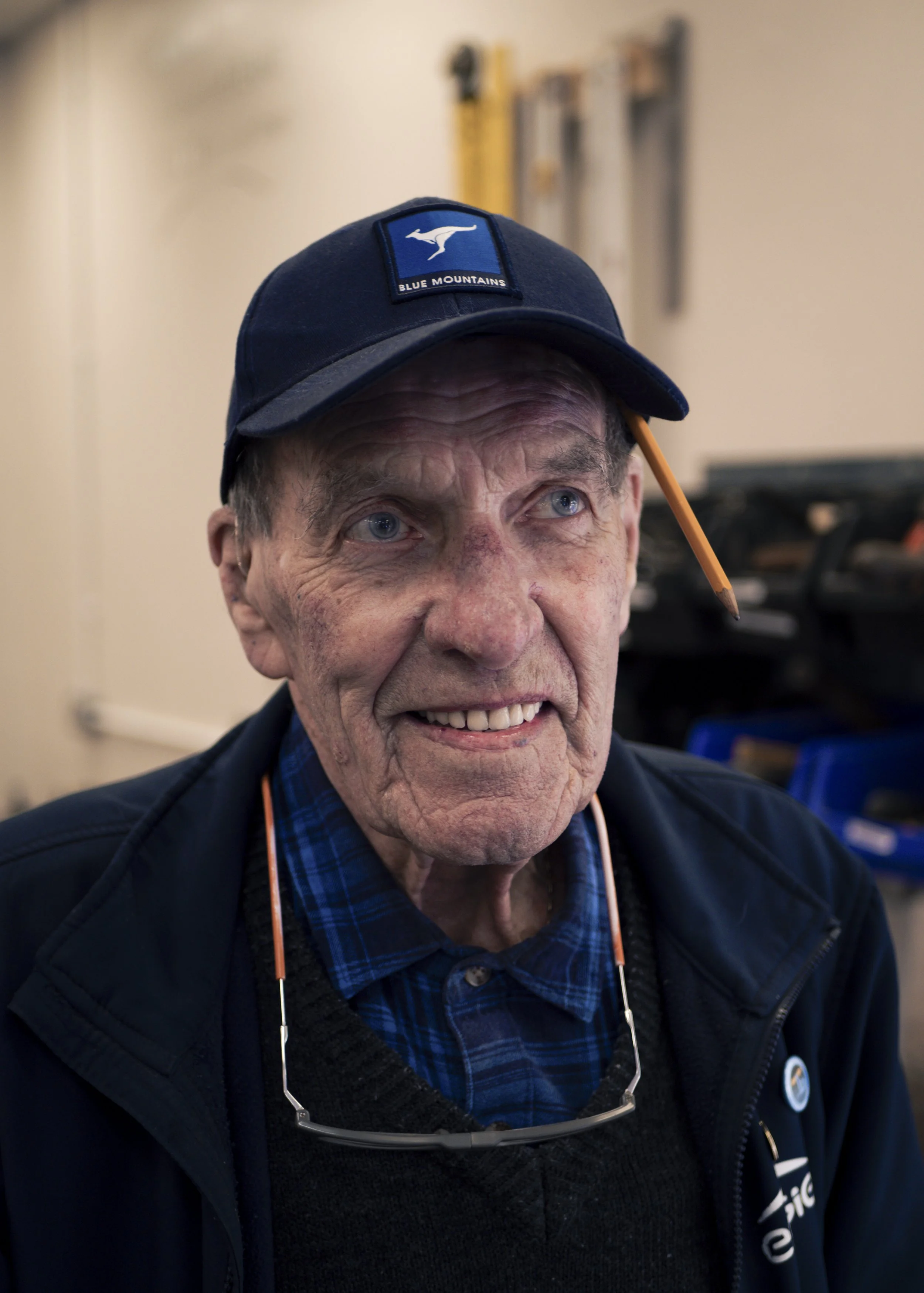 Close-up of an elderly man with blue eyes, wearing a navy baseball cap with a kangaroo logo and the words 'Blue Mountains,' a dark jacket, and glasses hanging around his neck, indoors with tools or equipment in the background.