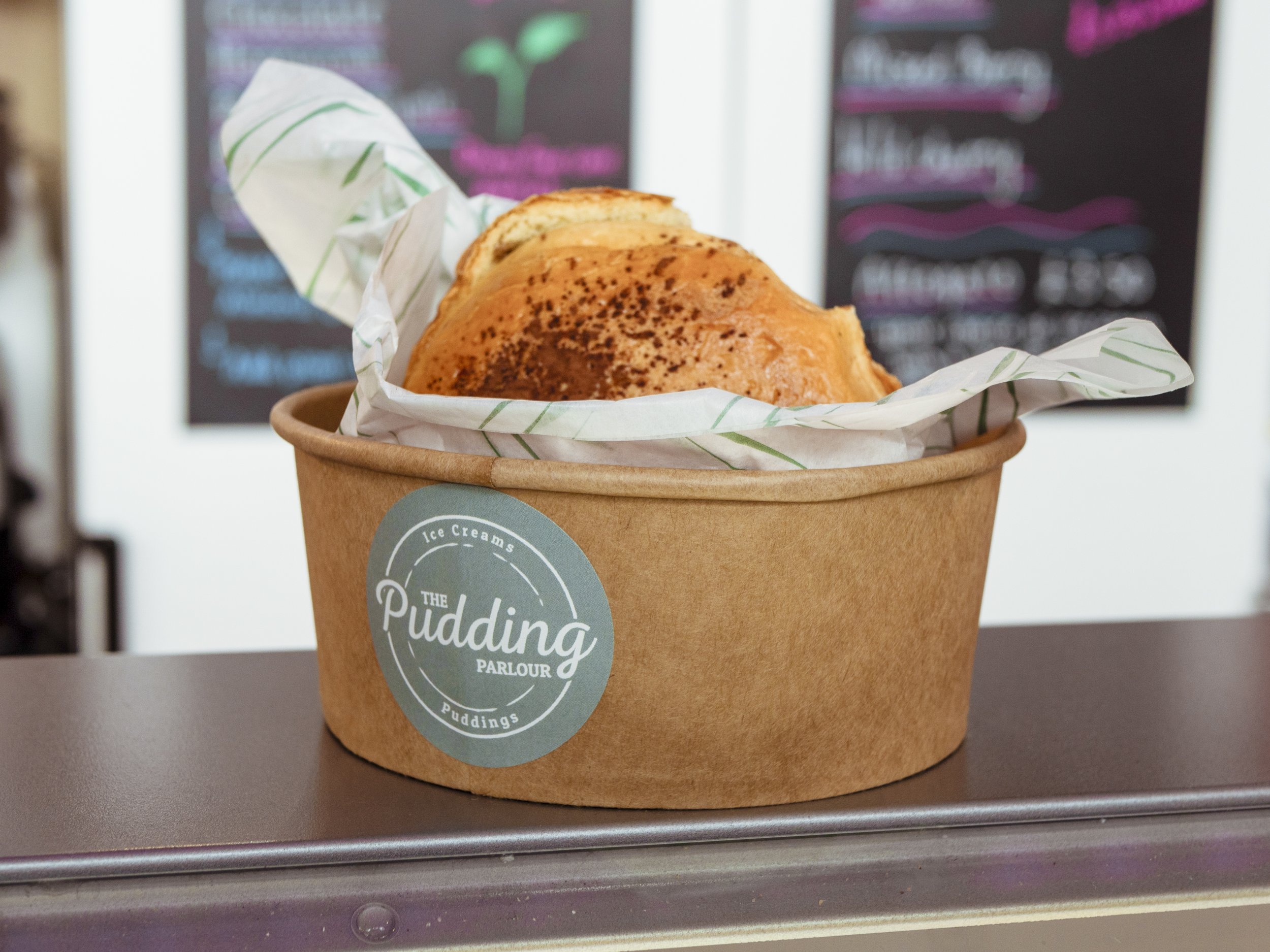 A round paper container with a sticker that reads 'The Pudding Parlour' contains a large serving of bread pudding with a crispy top, lined with parchment paper, placed on a metal surface.
