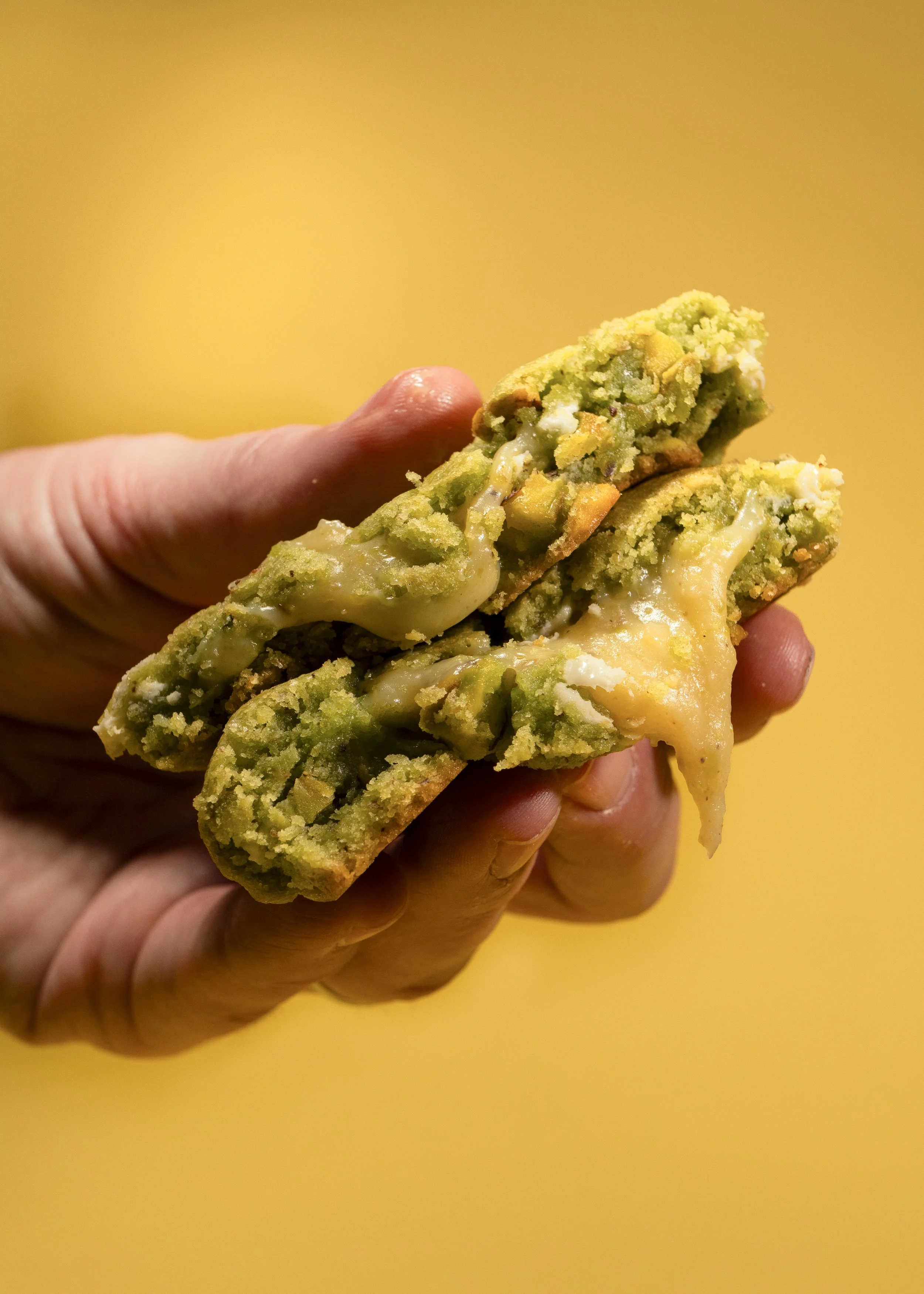 Close-up of a person's hand holding a piece of green chocolate chip cookie with melted cheese.