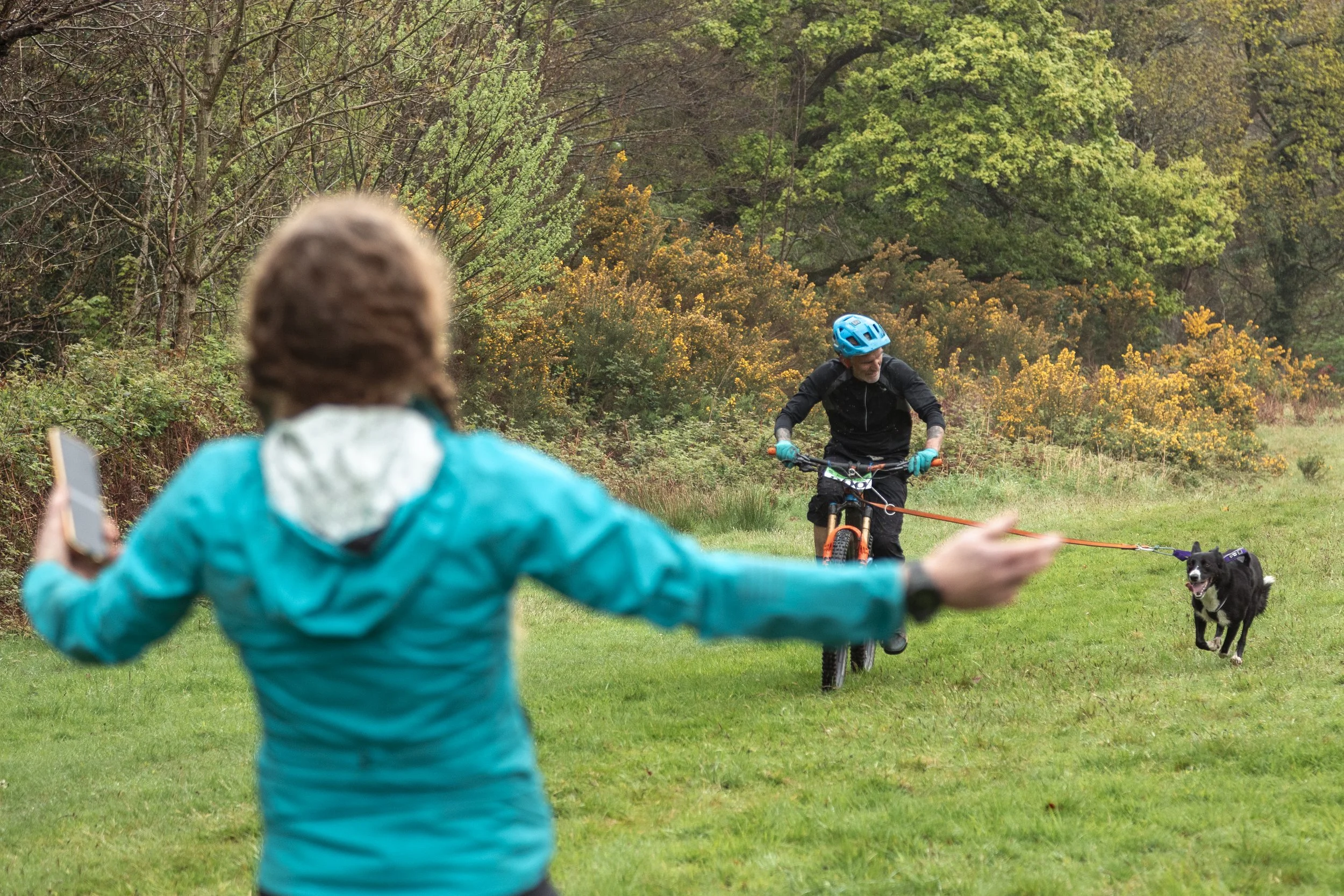 A person in a blue jacket is holding a phone with their left hand while reaching out with their right hand. In the background, a cyclist wearing a blue helmet and black clothing rides a bike with a dog on a slackline. The dog appears to be balancing 