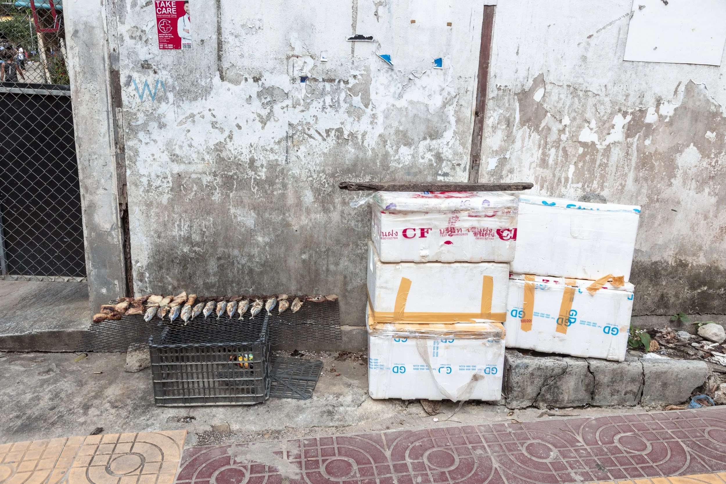A street scene with three large white styrofoam boxes stacked against a weathered, peeling wall, and a small grill with fish drying on it to the left.