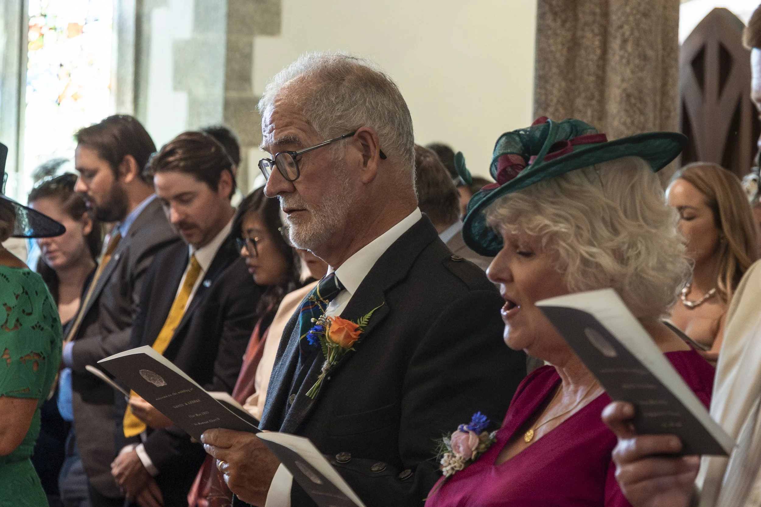 People participating in a religious ceremony inside a church, holding prayer books, dressed in formal attire with some women wearing decorative hats.