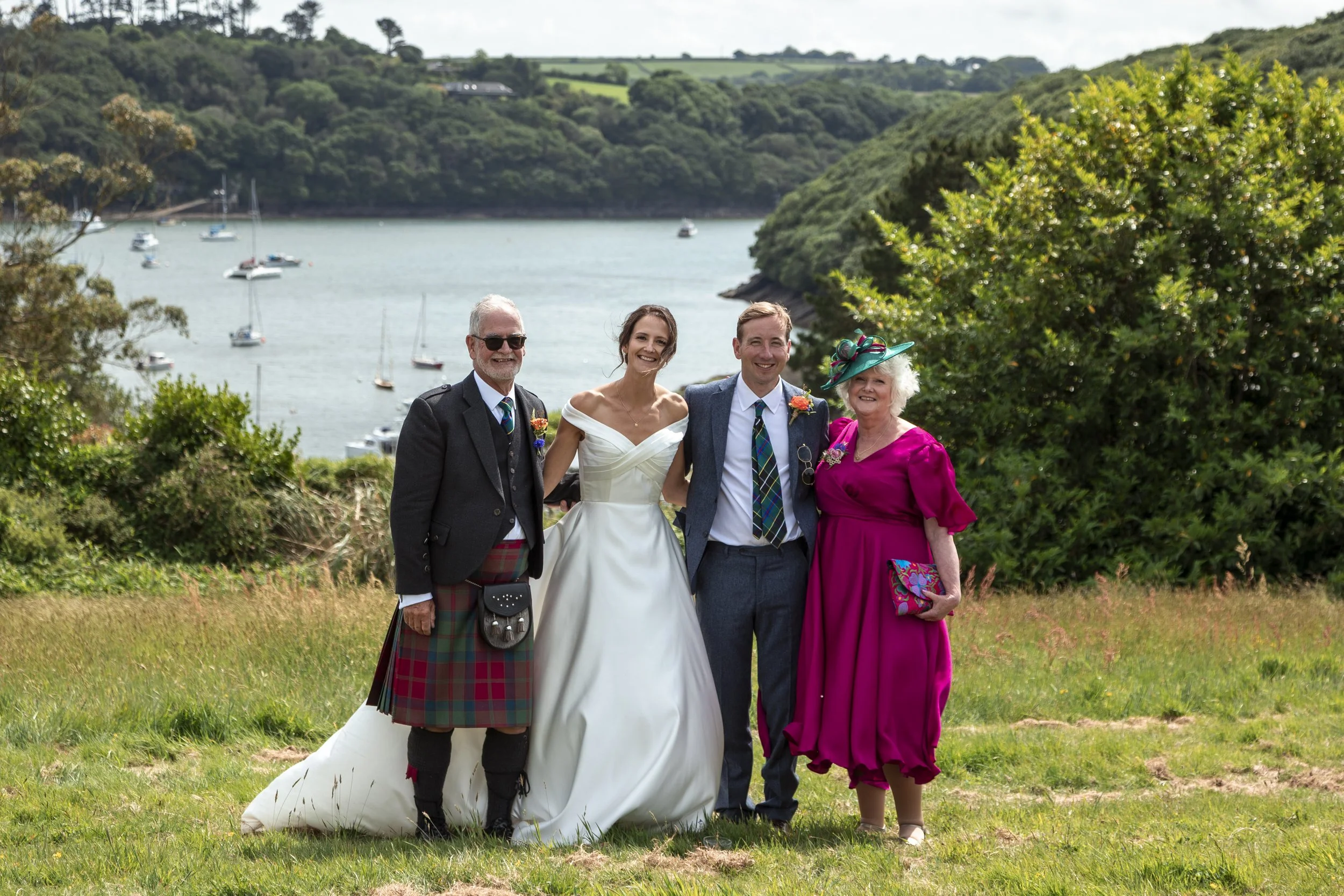 A group of four people, including a bride in a white wedding dress, standing outdoors on a grassy area near a coastline with boats anchored in the water and green hills in the background.