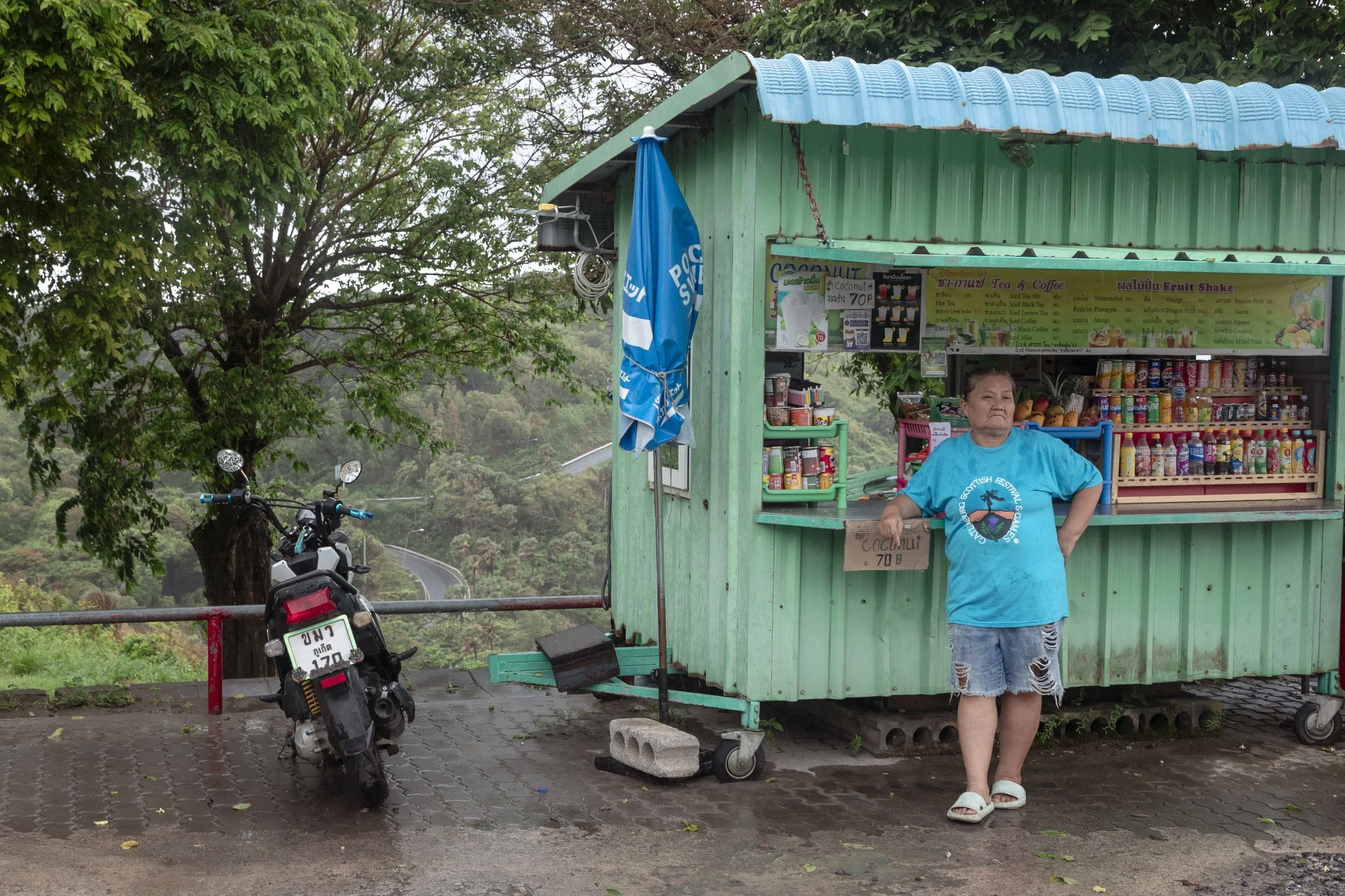 A woman in a blue T-shirt and ripped denim shorts stands outside a small green roadside shop with snacks and drinks, next to a parked motorcycle, on a wet, paved surface with trees in the background.