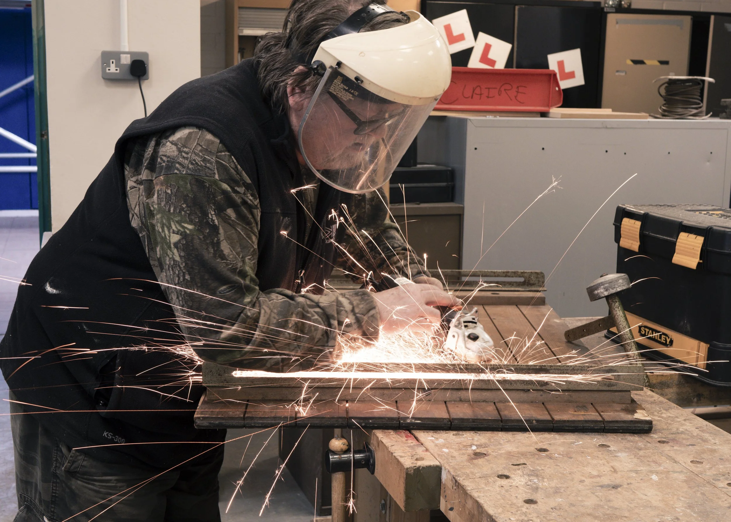 A person wearing a protective helmet and camouflage jacket is welding metal piece on a workbench, with sparks flying around.
