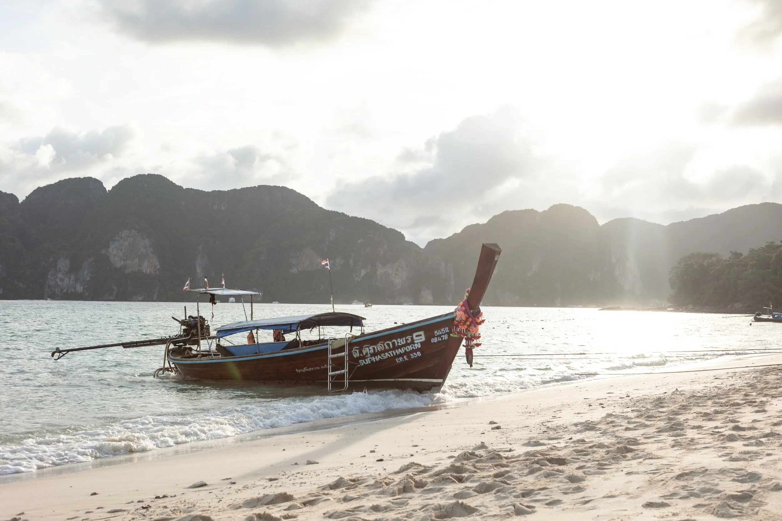 A traditional Thai longtail boat floating near the sandy beach with mountains in the background and the sun setting.