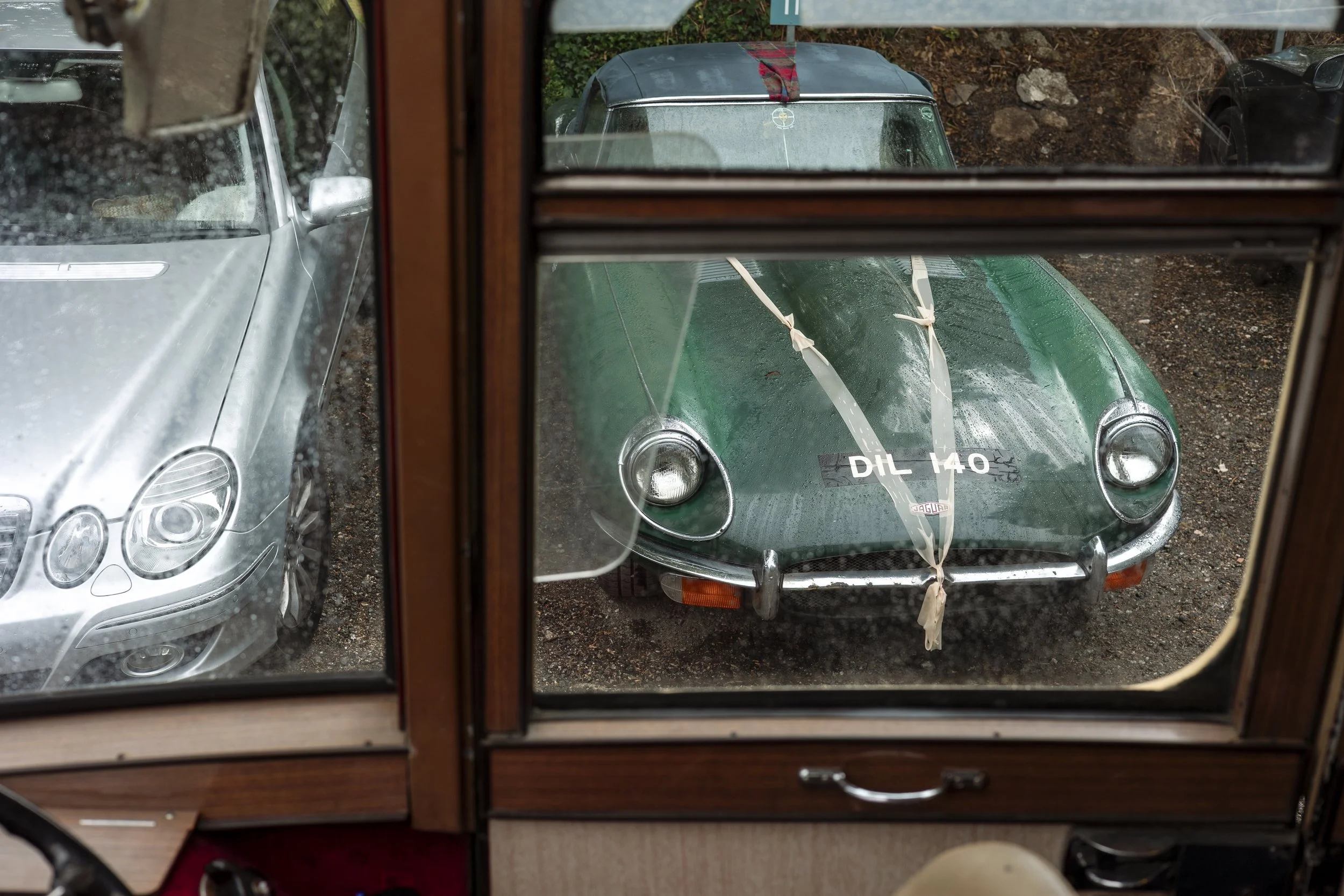 View through a window showing three parked cars outside, including a classic green vintage sports car with a license plate that reads 'DIL 140' and straps on its hood, surrounded by a gravel ground.