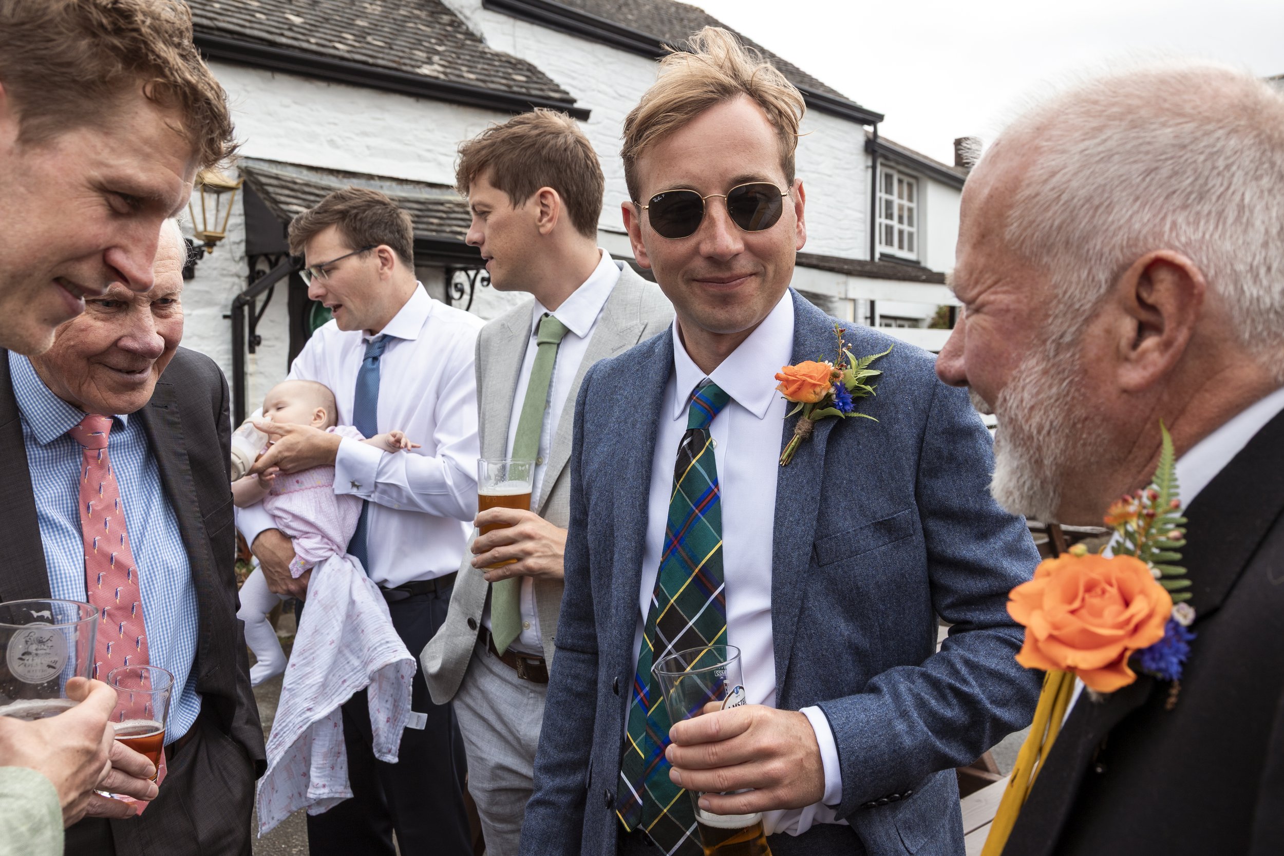 Group of men at an outdoor social event, dressed in suits with colorful ties and boutonnieres, holding drinks and engaging in conversation.