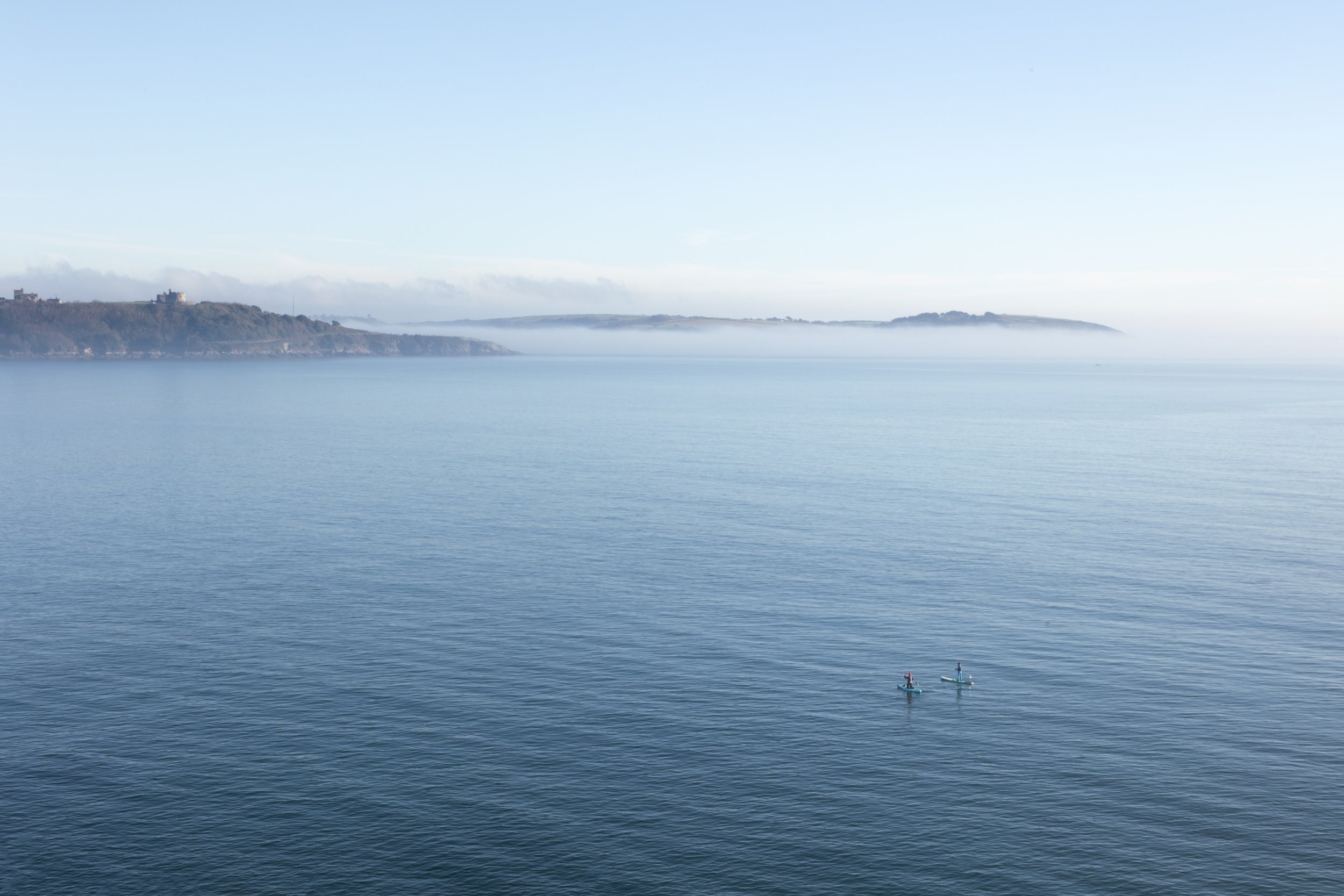 A calm body of water with two paddleboarders. Distant land and fog over the hills in the background.