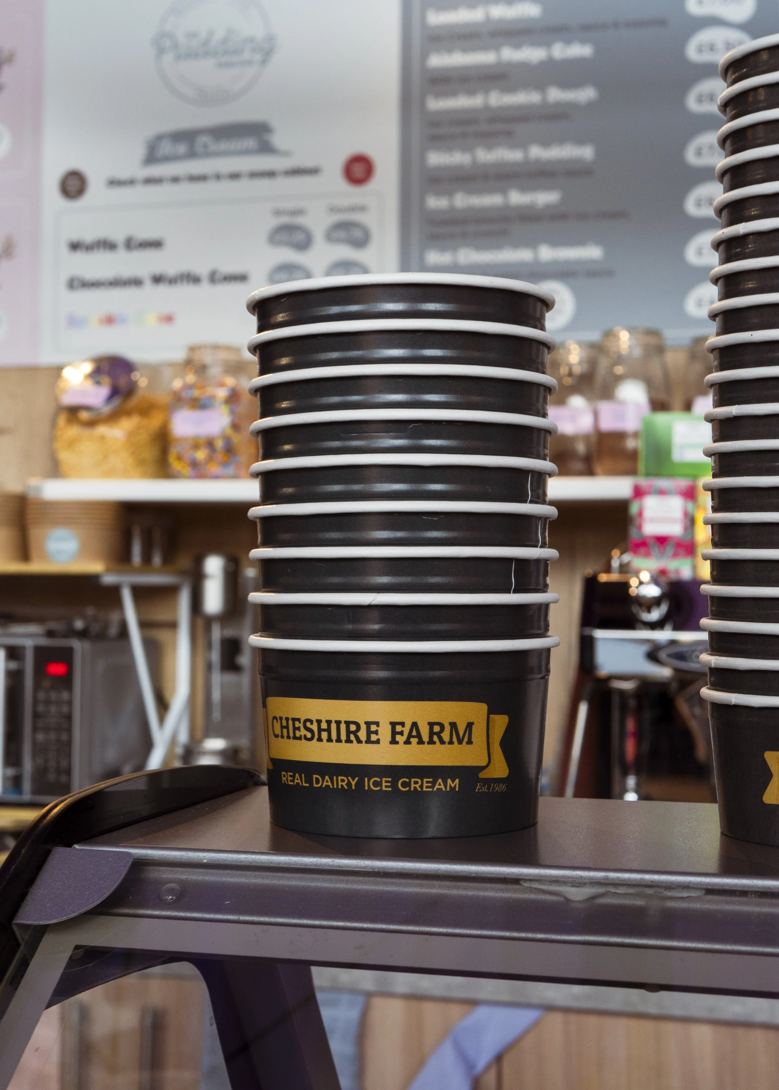 Stack of black paper cups with Chester Farm logo and yellow label, on a table in a cafe or ice cream shop, with a blurry background of menu and jars of toppings.