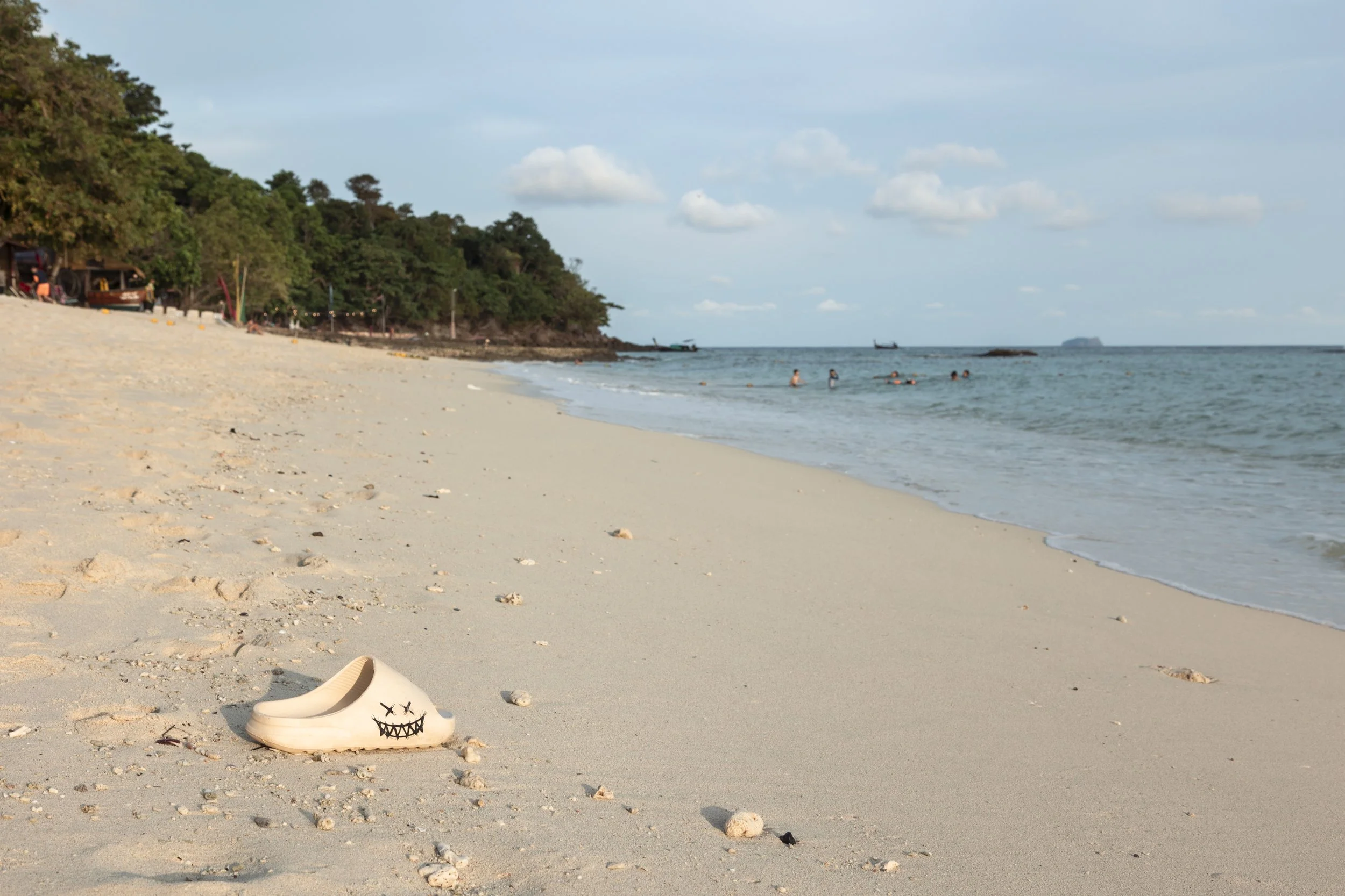 A sandy beach with a sea shoe painted with a face drawing in the foreground, trees and huts on the left, and people swimming in the ocean.