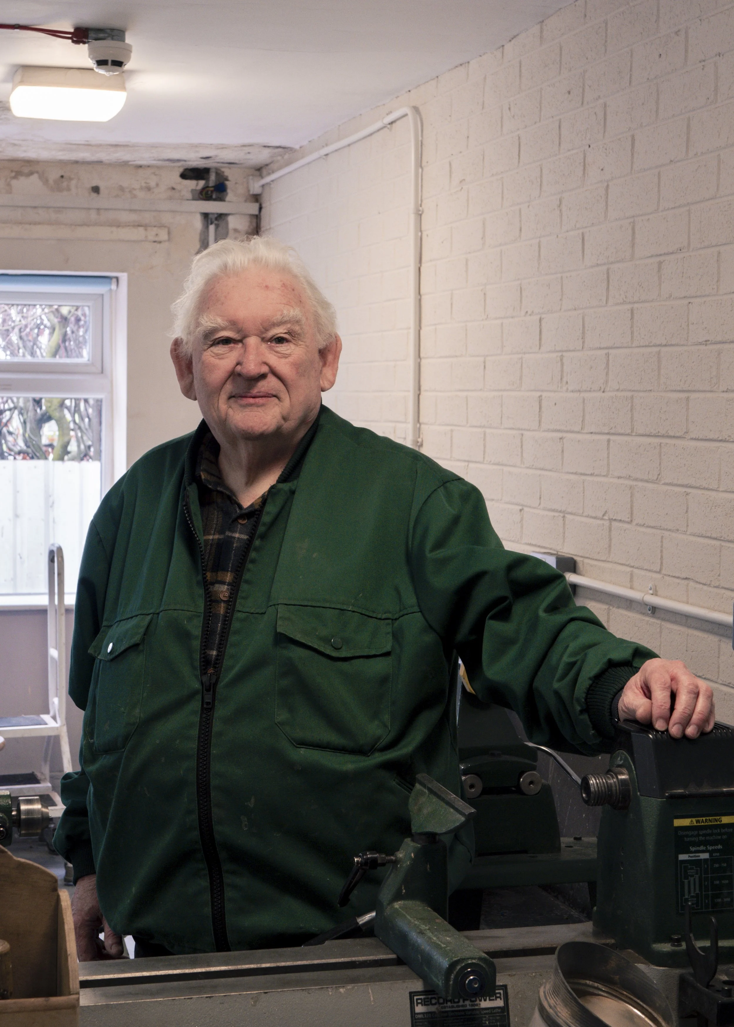 An elderly man with white hair wearing a green jacket standing in a workshop or garage with tools, near a white brick wall and window.