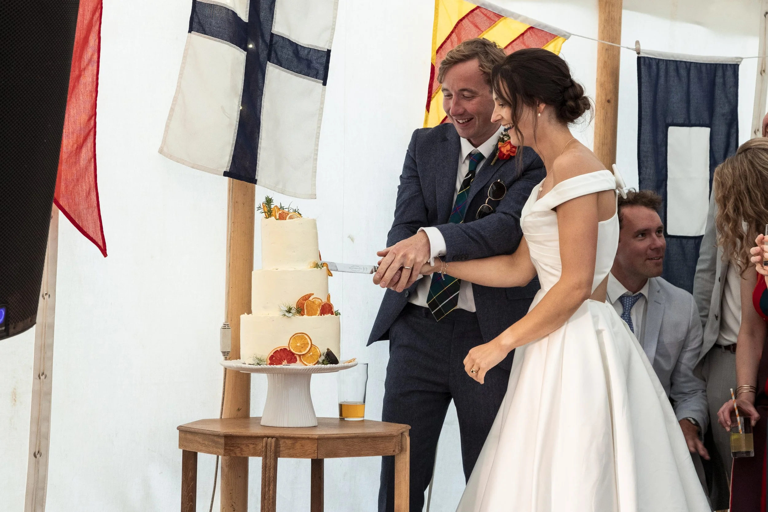 Bride and groom cut a wedding cake decorated with orange and grapefruit slices under Union Jack flags at their wedding celebration.
