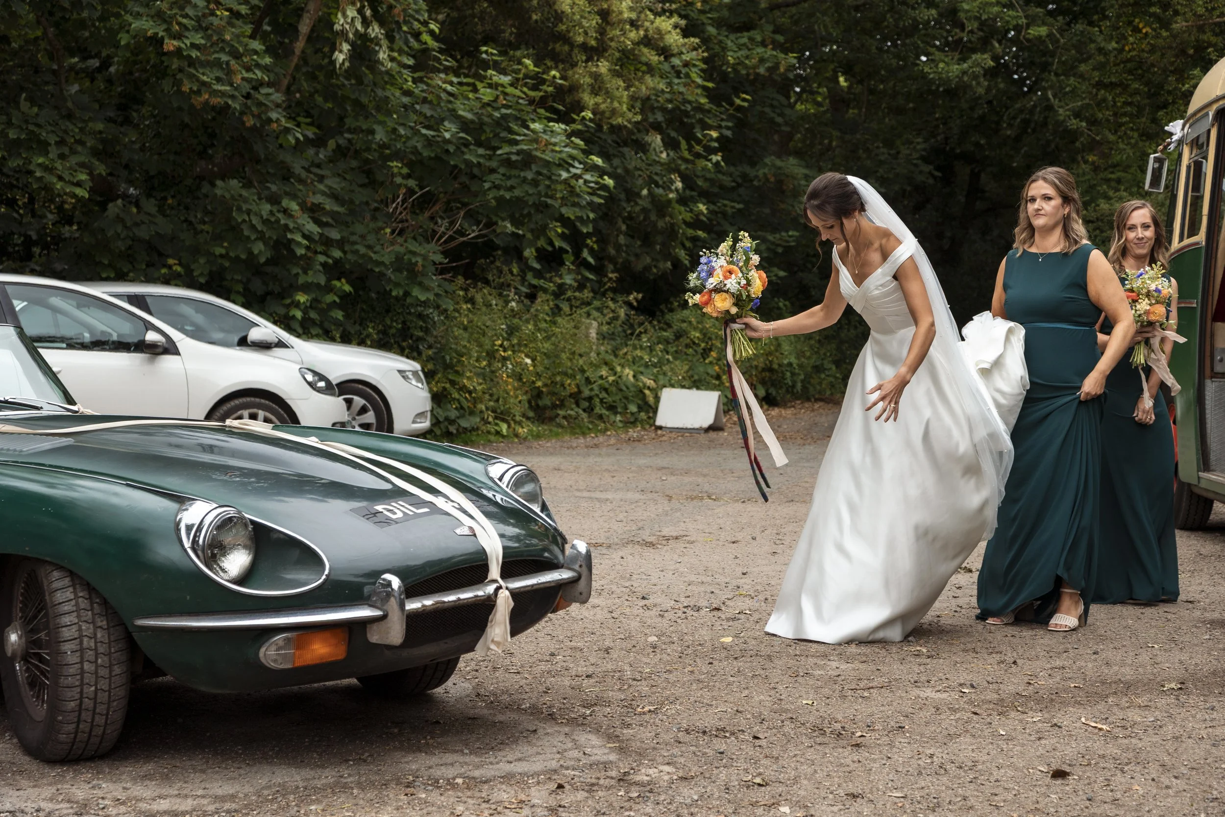 A bride in a white wedding dress and veil holding a bouquet, approaches a vintage green racing car with white ribbon and a bow tied around the front. Two bridesmaids in teal dresses follow her, each holding bouquets, as they stand near a bus or troll