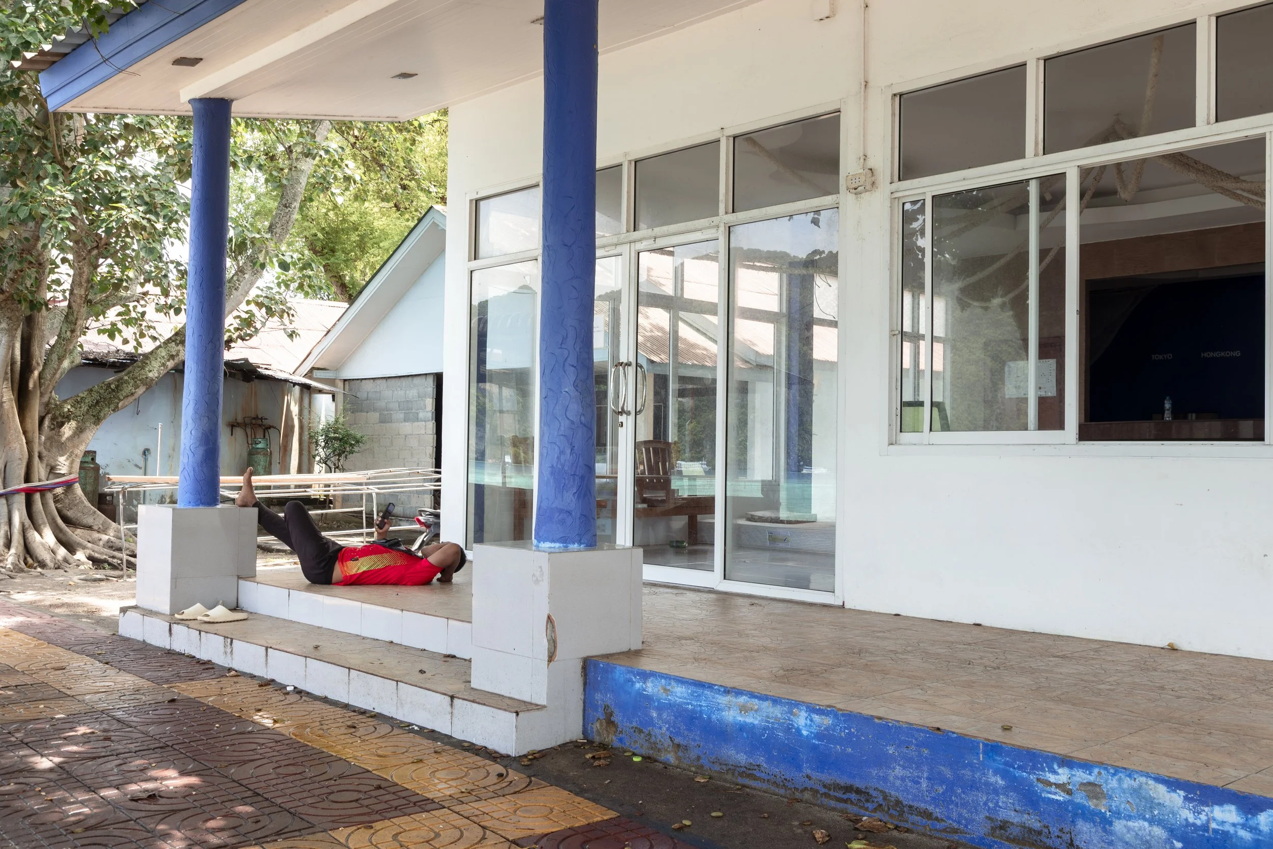 A person lying on a small tiled porch area outside a building, wearing a red shirt and black pants, looking at a phone. The porch is elevated with three steps down to a brick sidewalk. The building has large windows and two blue columns supporting th