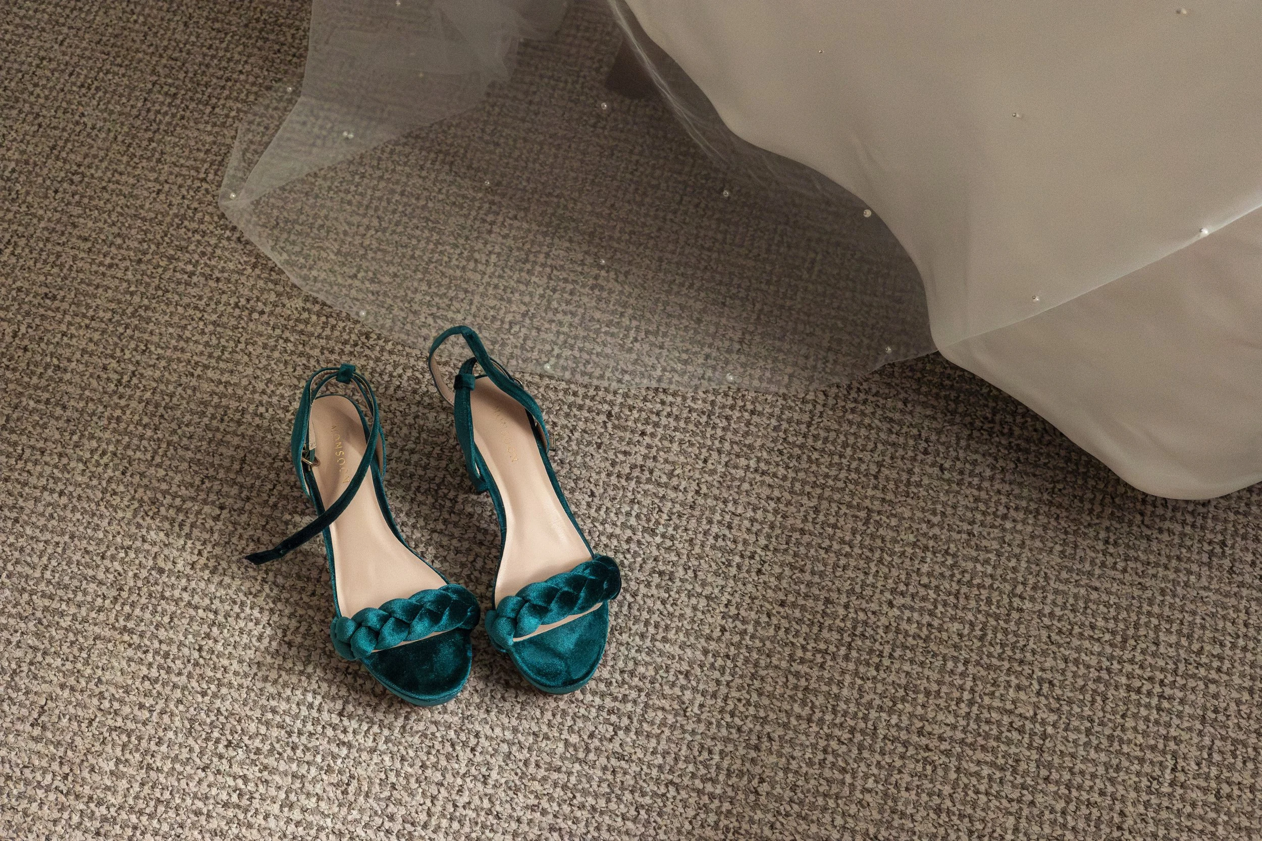Pair of teal high heel shoes with braided strap on a beige carpeted floor near a bed with a white bedspread and clear plastic canopy.