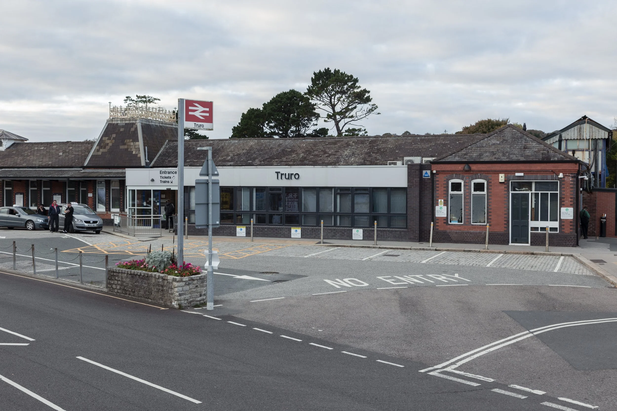 Exterior of Truro train station with a platform sign, parking lot, and a small brick building, under a cloudy sky.