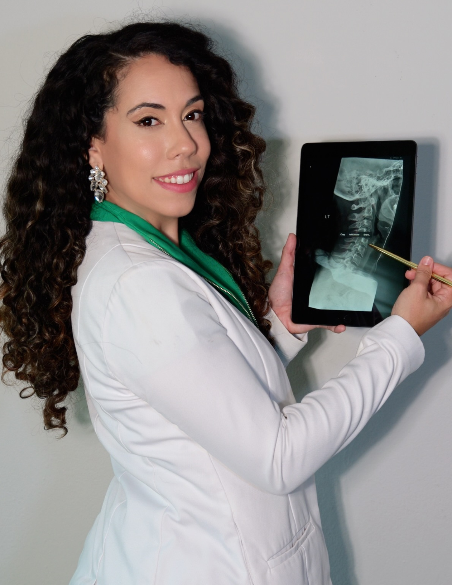 A smiling female doctor or Chiropractic (Dr. Trinidad) with curly hair  wearing a white coat and green scrubs, holding a tablet displaying an X-ray image of a neck and upper spine, pointing to the X-ray with a gold pen.