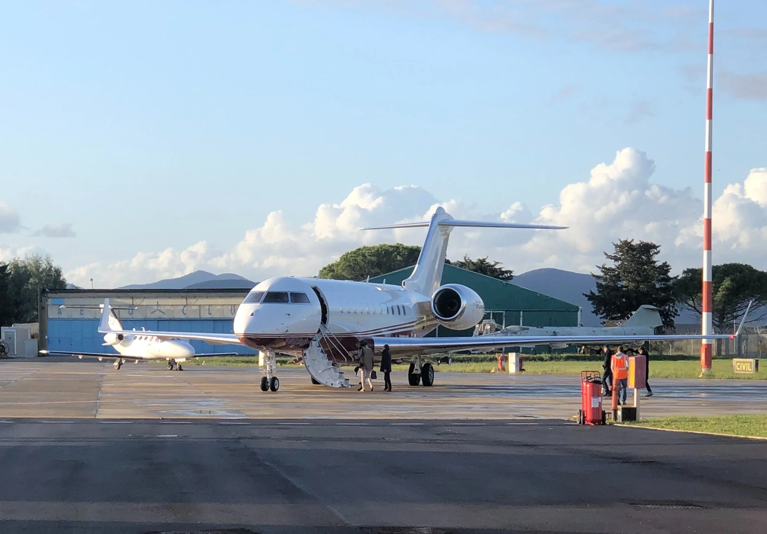 Private jet on a tarmac with people boarding, mountains and clouds in the background.