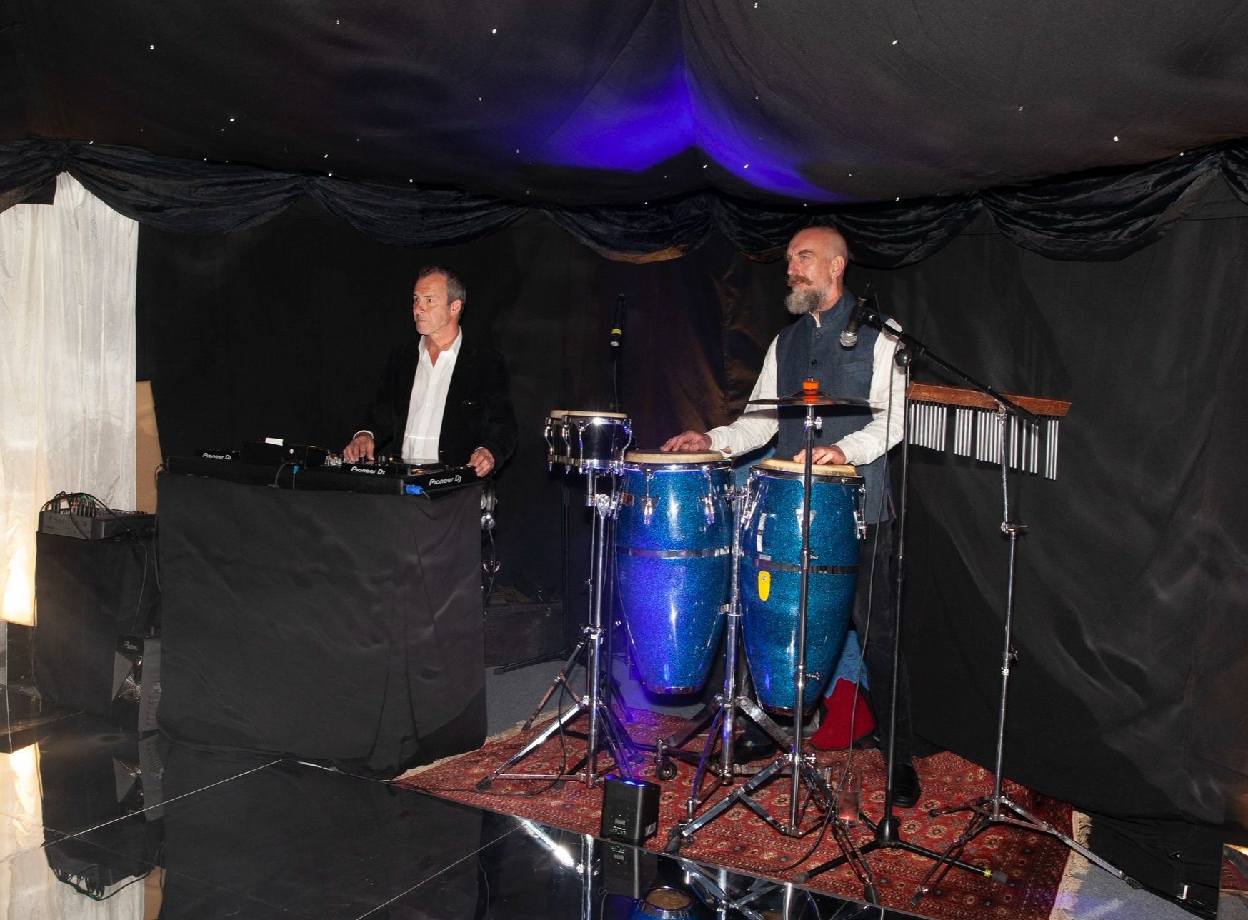 Two musicians, one at a DJ booth and the other playing conga drums, perform on a stage with black curtains. The setup includes a microphone and percussion instruments, with a reflective floor and overhead purple lighting.