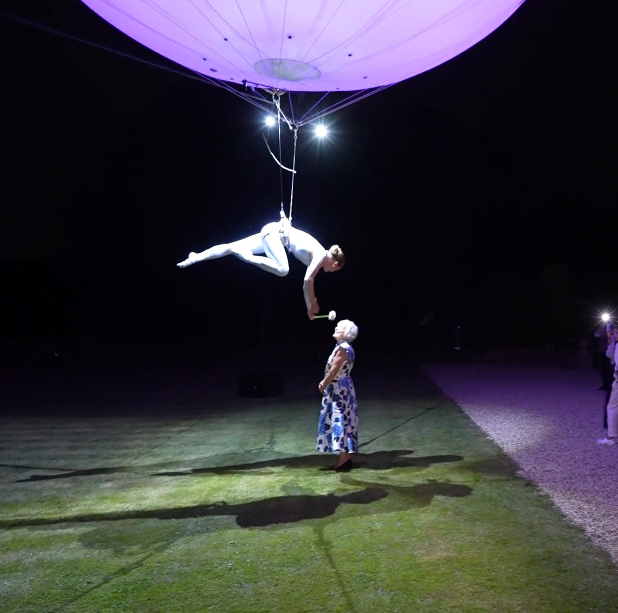 Aerial performer suspended from an illuminated balloon, holding a flower towards an elderly woman wearing a floral dress.