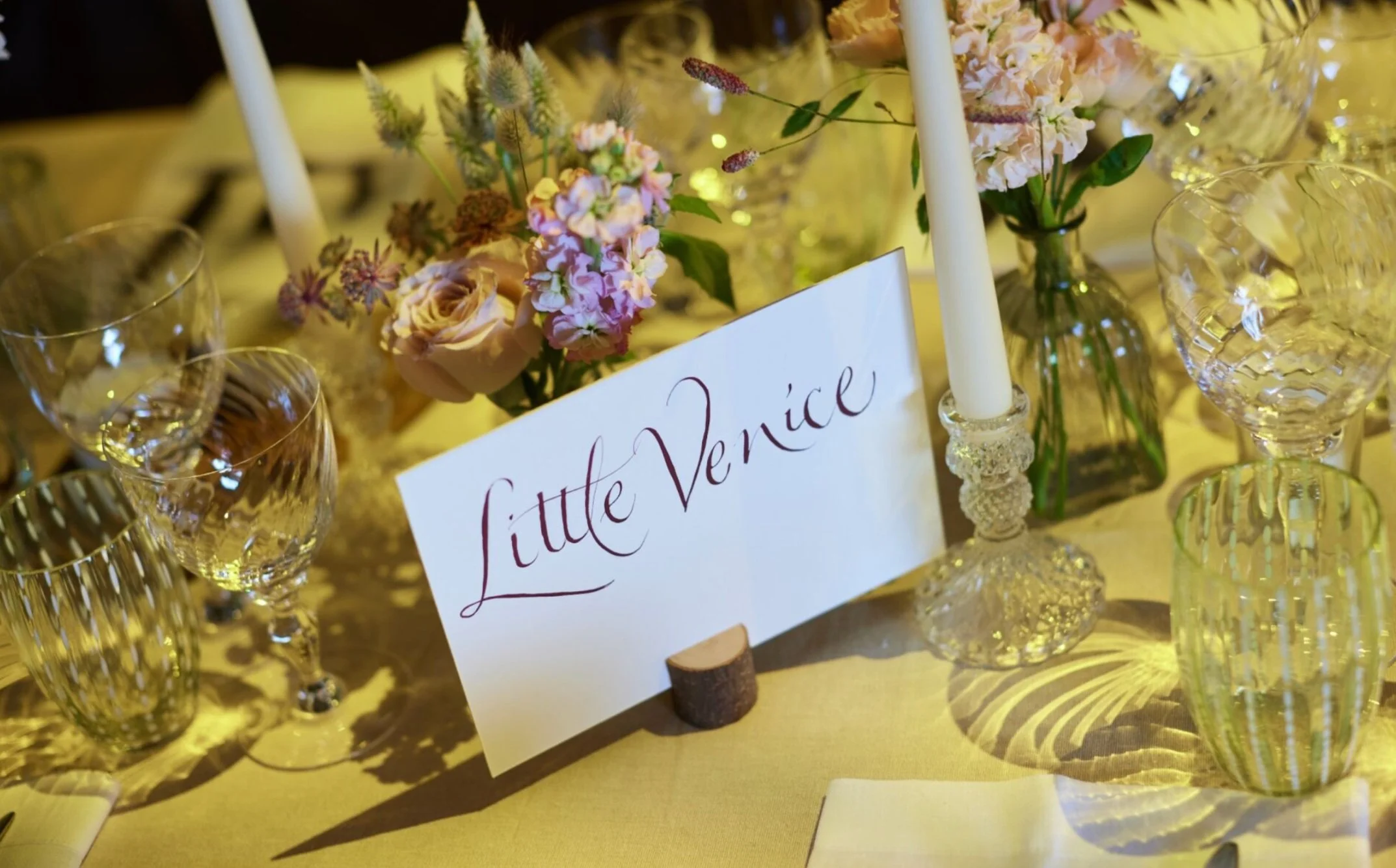 Table setting with handwritten card reading 'Little Venice', surrounded by glasses, flowers, and candles.