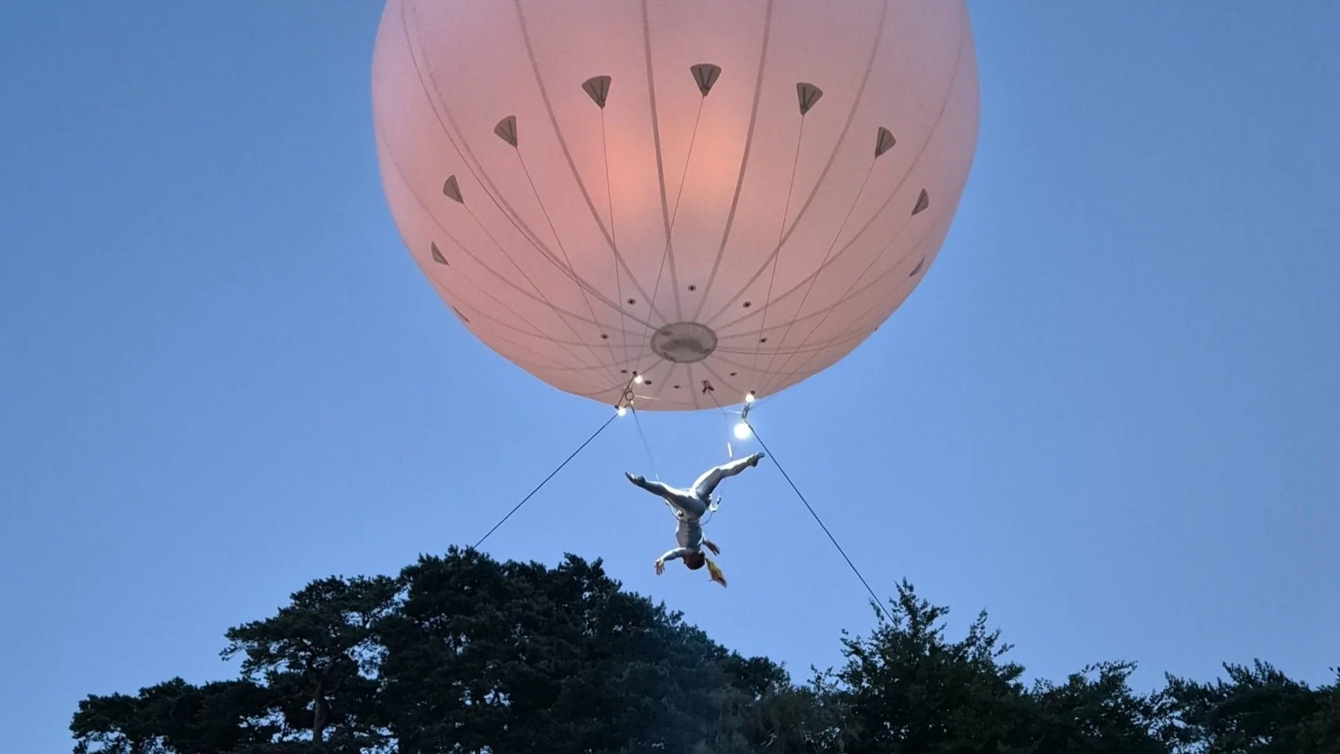 Large balloon with an acrobat suspended beneath, performing an aerial act against a twilight sky.