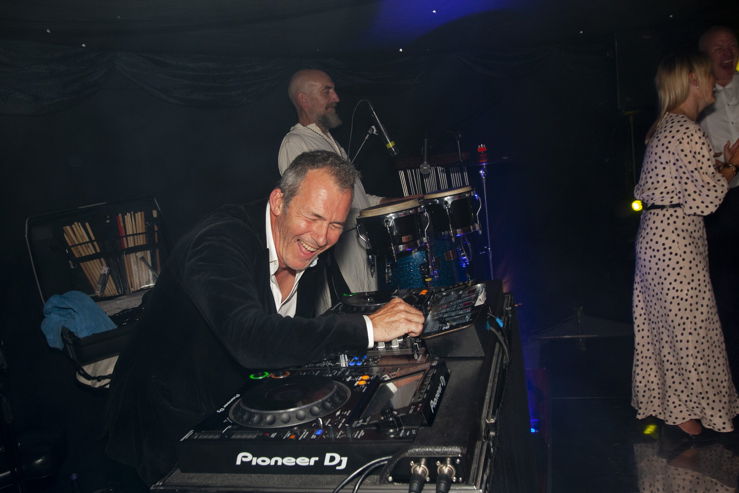 DJ and musician performing at an event with people dancing and smiling in the background.