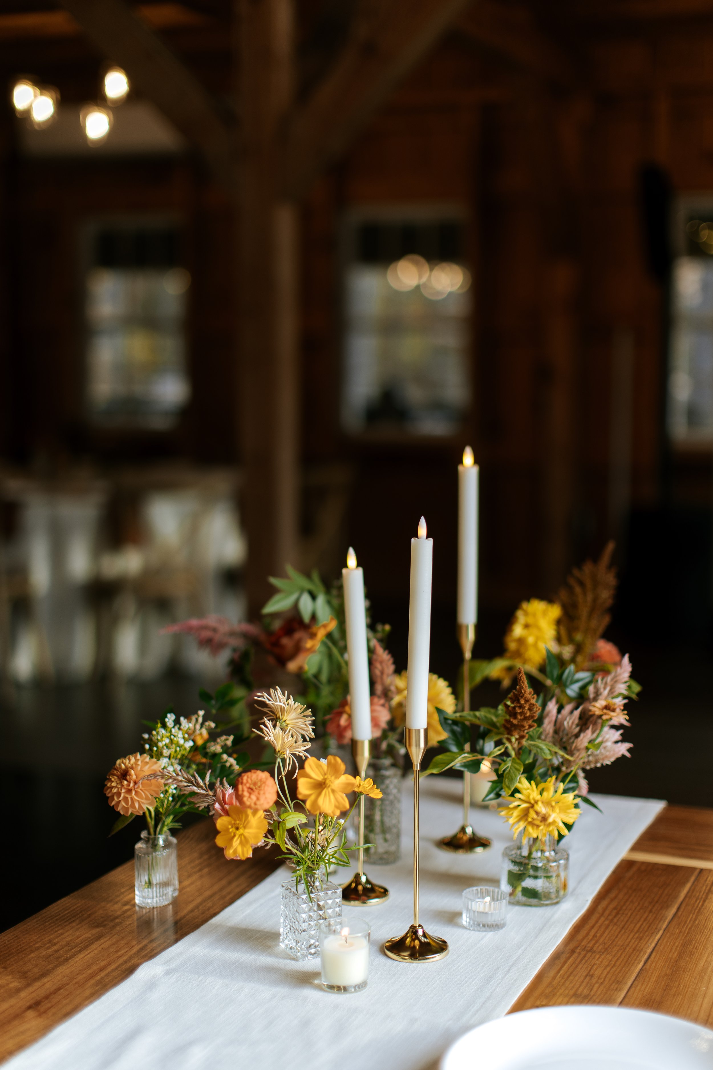 Table decorated with candles and colorful flowers in small glass vases inside a rustic wooden venue.