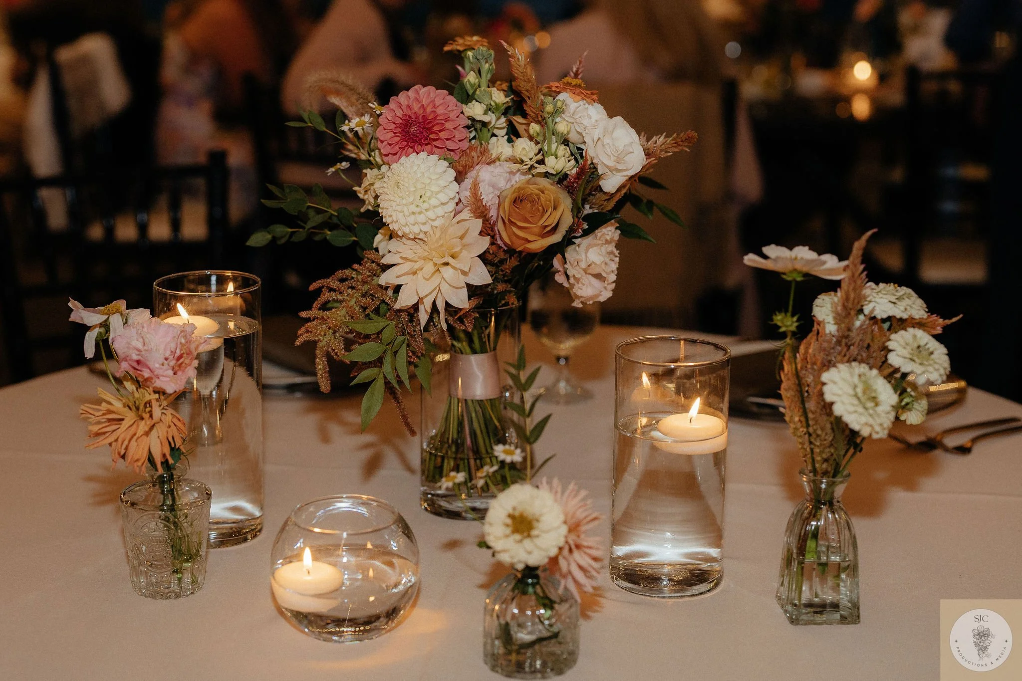 Elegant dinner table with floral centerpiece and floating candles in glass holders, featuring roses and dahlias in soft pastel tones at a dimly lit event.