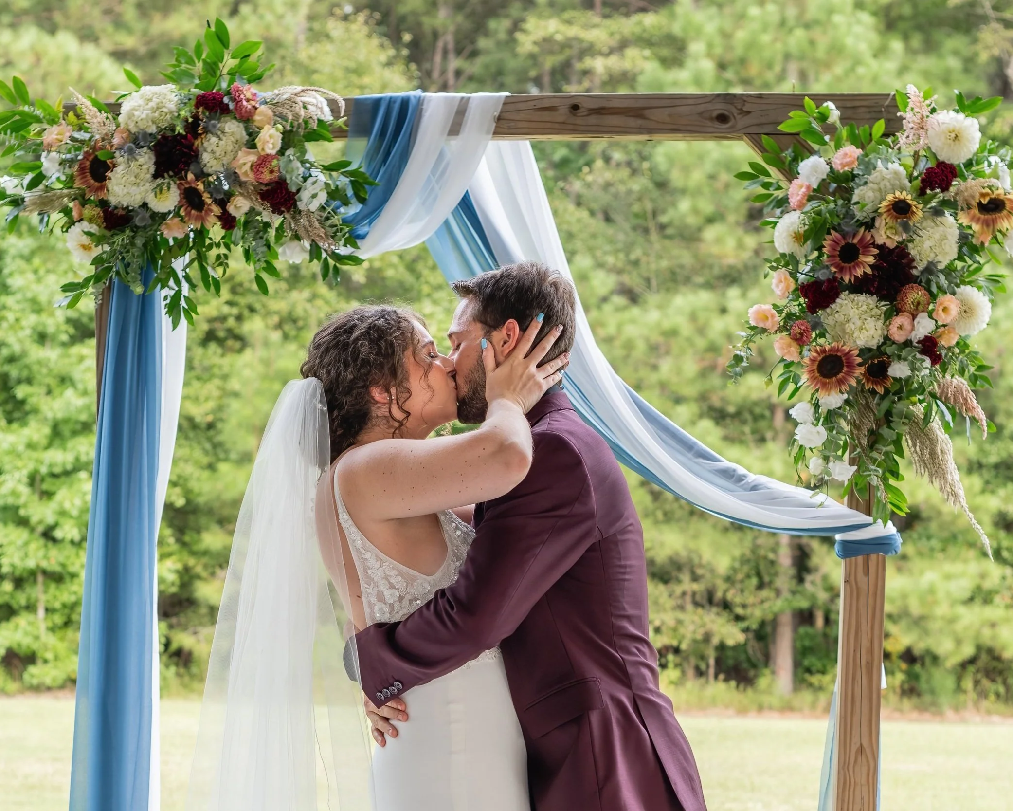 A bride and groom kiss under a wooden wedding arch decorated with flowers and draped fabric outdoors during daytime.