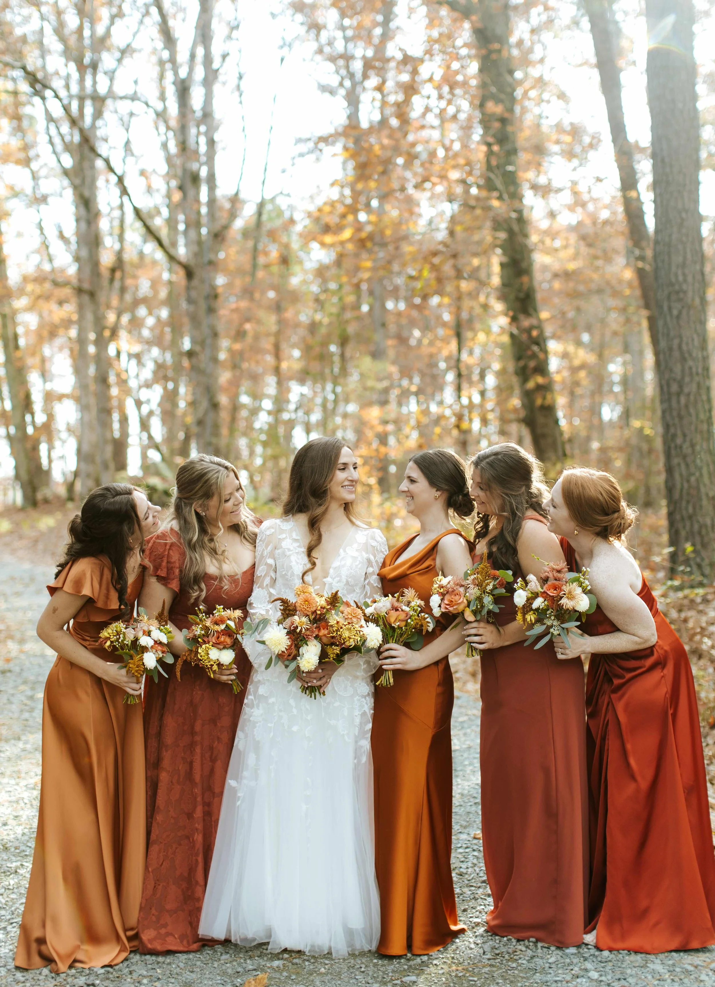 Bride in white dress with five bridesmaids in rust-colored dresses holding bouquets, standing outdoors in a wooded area with autumn leaves.