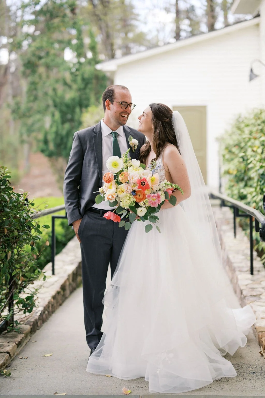 A bride and groom laugh together with her early spring citrus pink wedding bouquet at the Parlour at Mann's Chapel in Chapel Hill, NC