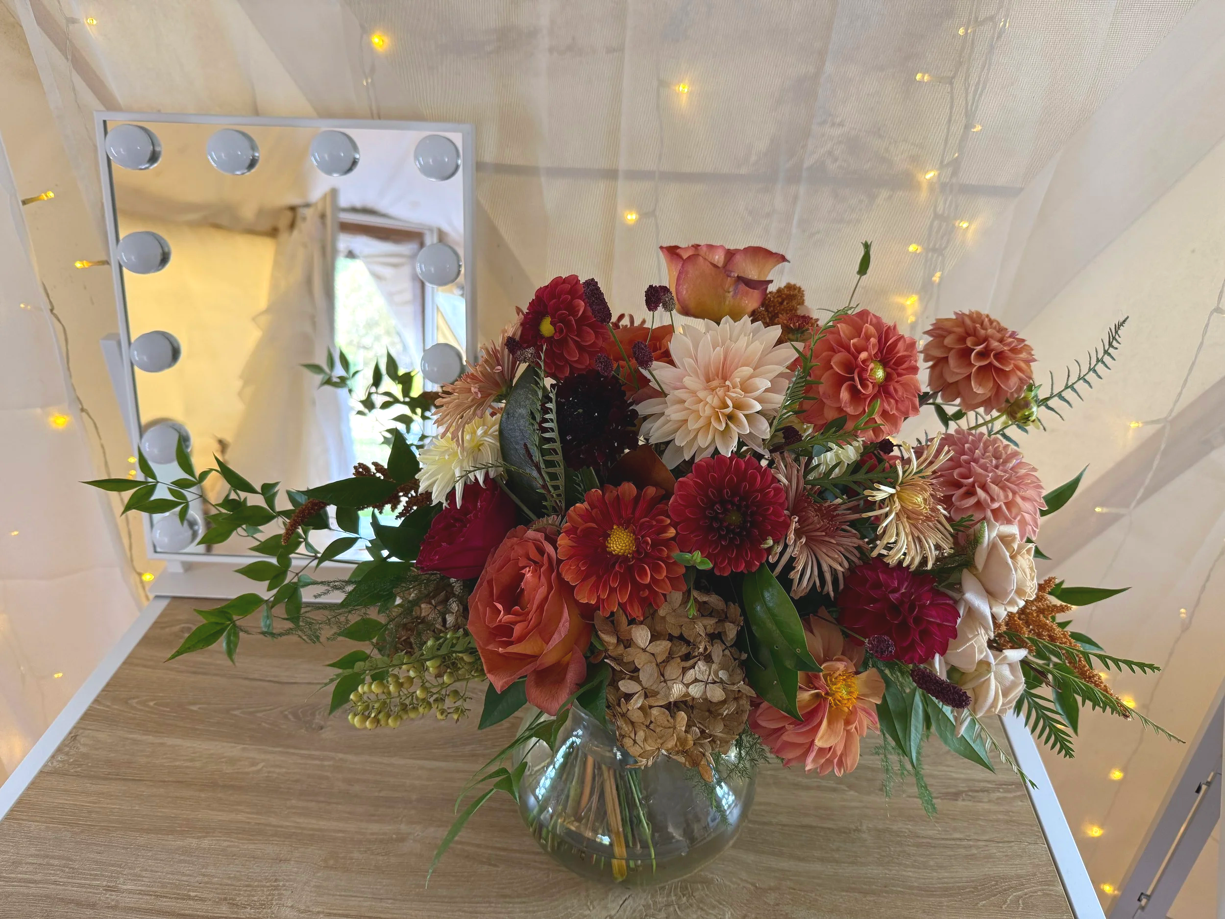A colorful bouquet of flowers in a glass vase placed on a wooden table, with a mirror reflecting a soft light and a background decorated with small yellow string lights.
