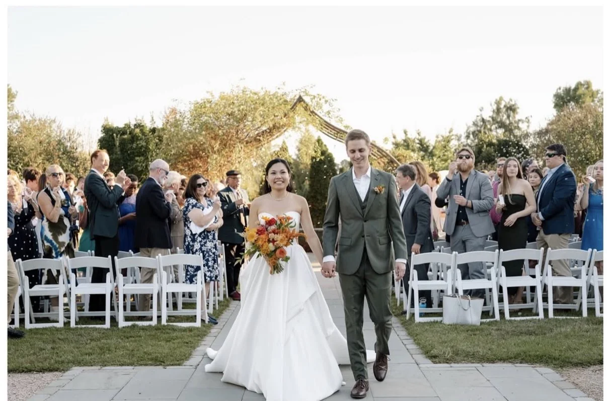 A newlywed couple walking hand in hand down the aisle at an outdoor wedding ceremony. The bride is wearing a strapless white wedding gown and holding a bouquet of orange, yellow, and red flowers. The groom is dressed in a gray suit with a white shirt and a boutonniere. Guests are clapping and watching the couple, with some taking photos.