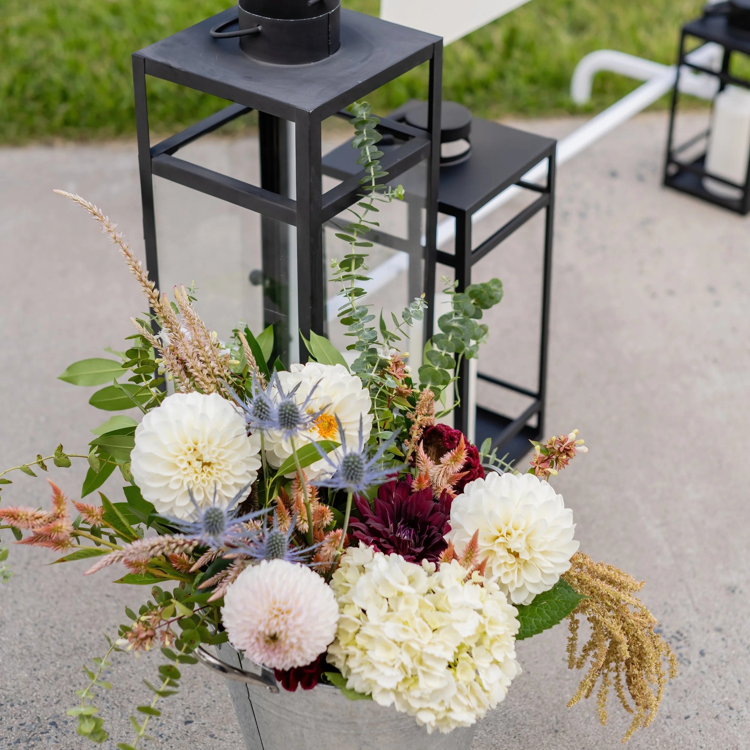 Vase filled with white, pink, burgundy, and lavender flowers and greenery, positioned near black lanterns on a concrete surface outside.