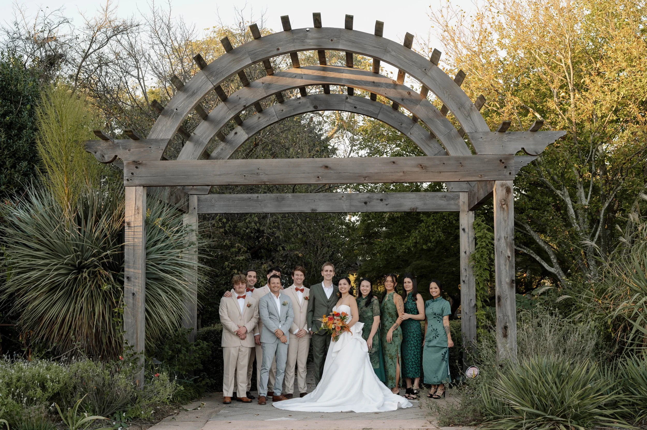 A fall wedding party poses beneath a wooden arbor at the JC Raulston Arboretum in Raleigh, NC