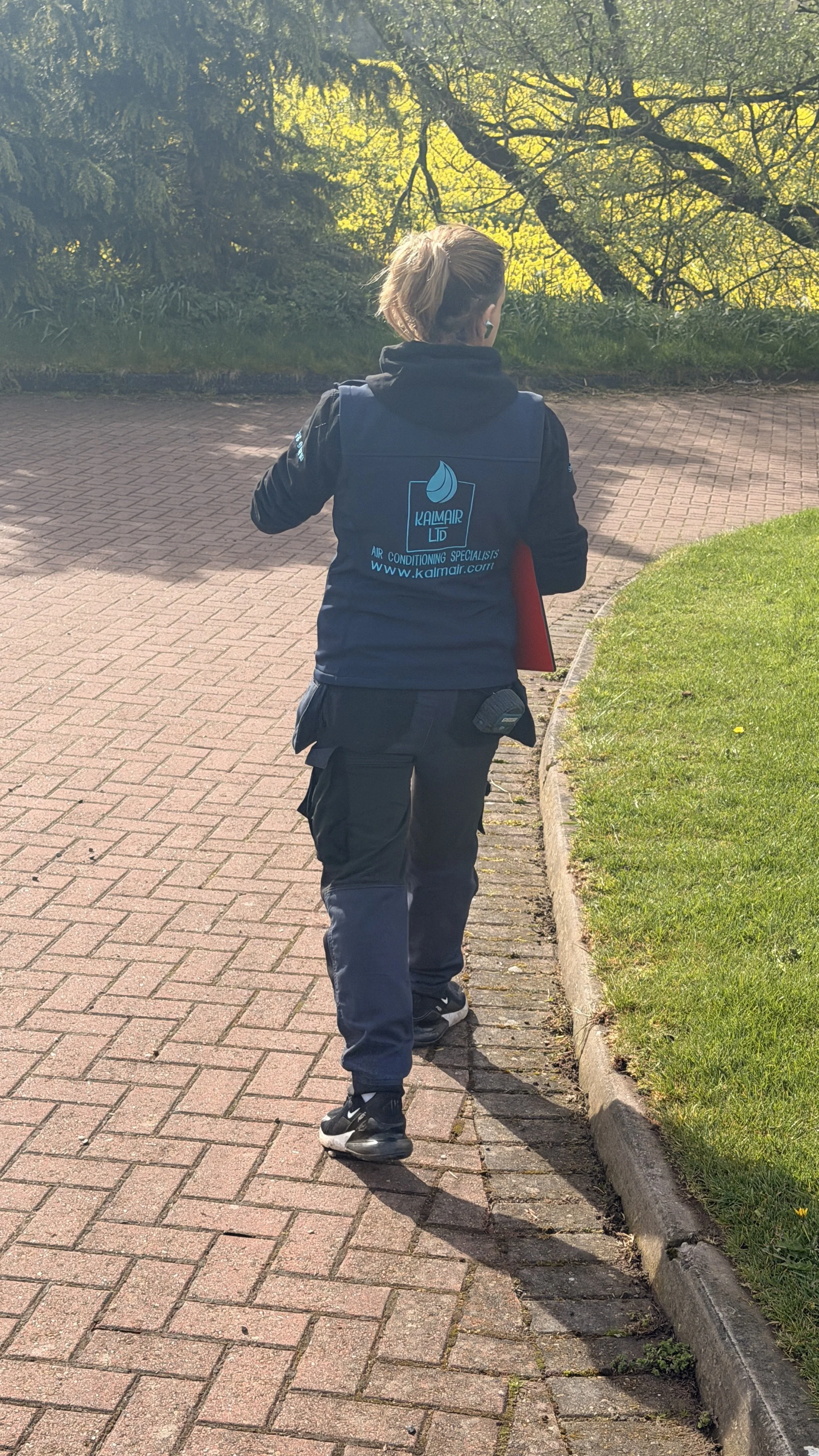 Smartly dressed professional person walking on a brick sidewalk near grass and trees with green foliage in the background. The person is wearing a KALMAIR LTD standard uniform.