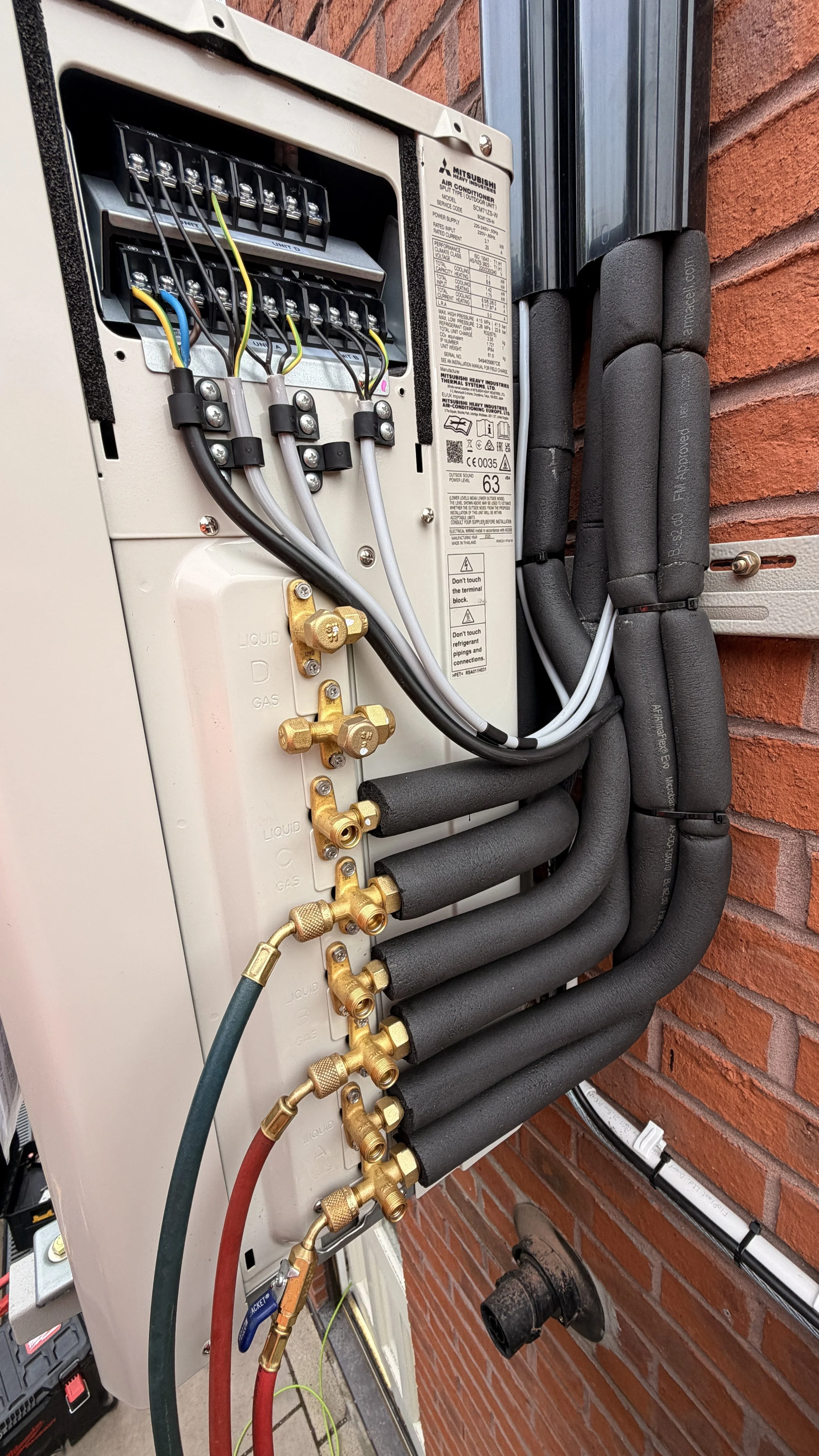 Close-up of an outdoor HVAC electrical and refrigerant connection panel mounted on a brick wall, with various pipes and wires connected.