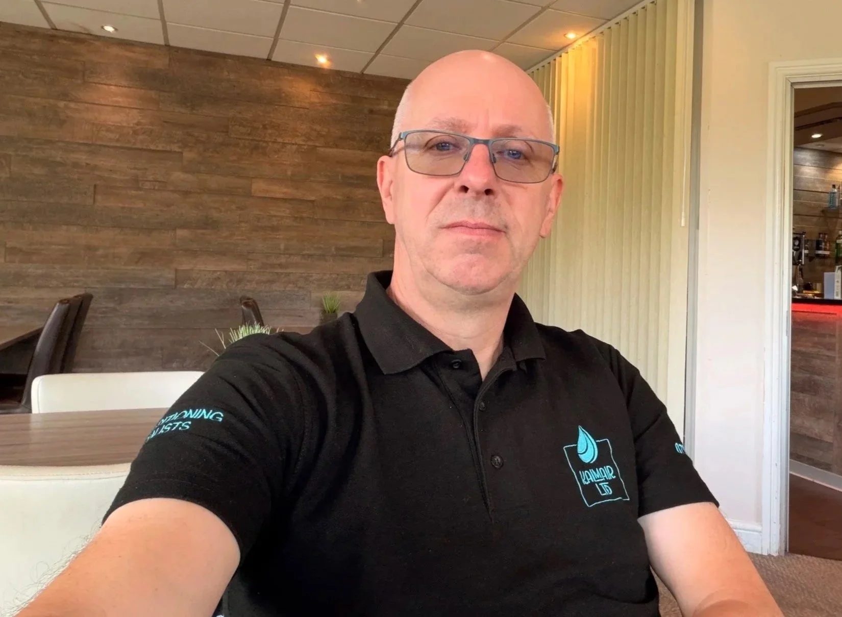 A bald man wearing glasses and a black shirt with a logo takes a selfie in a restaurant or cafe with wood-paneled walls and beige accents.
