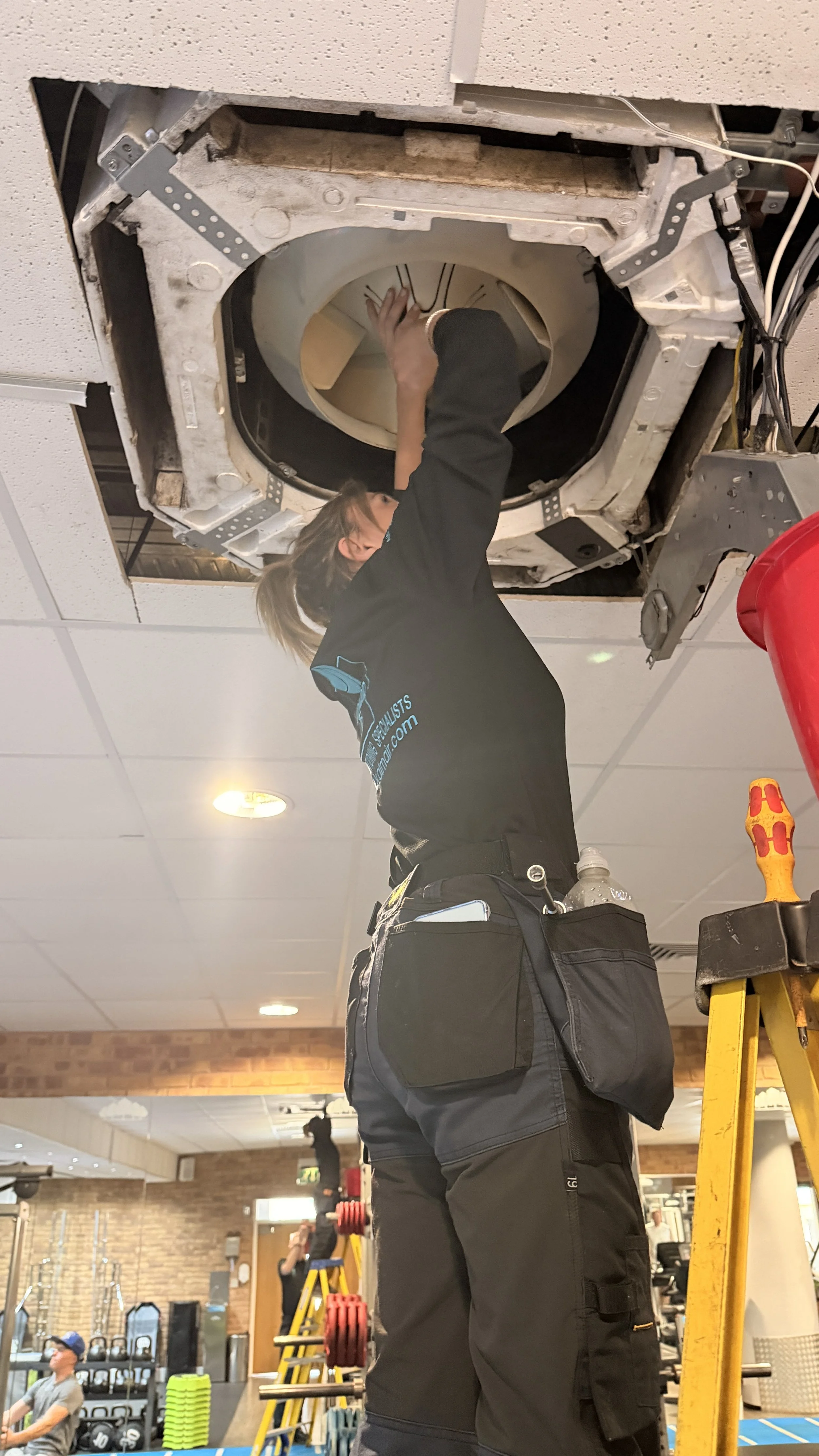A person in KALMAIR LTD standard uniform is repairing or inspecting an air conditioning unit in a gym or fitness center, with gym equipment and other people in the background.